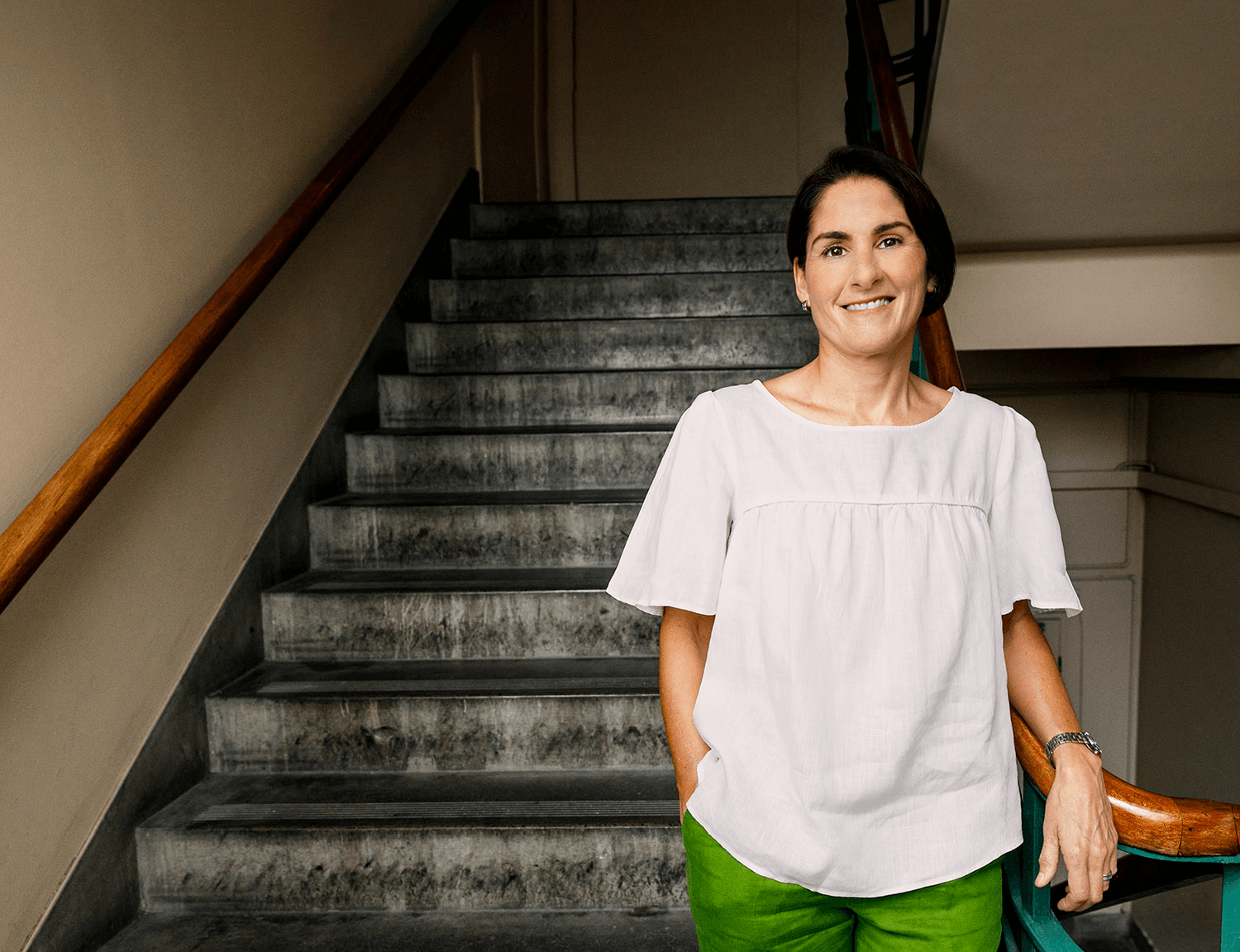 Woman in white blouse and green pants smiling, standing by staircase railing indoors.