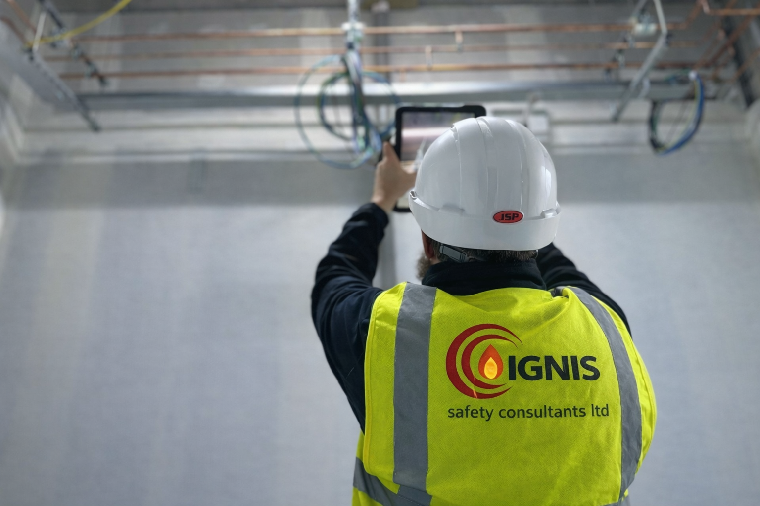 Worker wearing a white hard hat and yellow high-visibility vest labeled 'IGNIS safety consultants ltd' using a tablet to inspect electrical wiring.