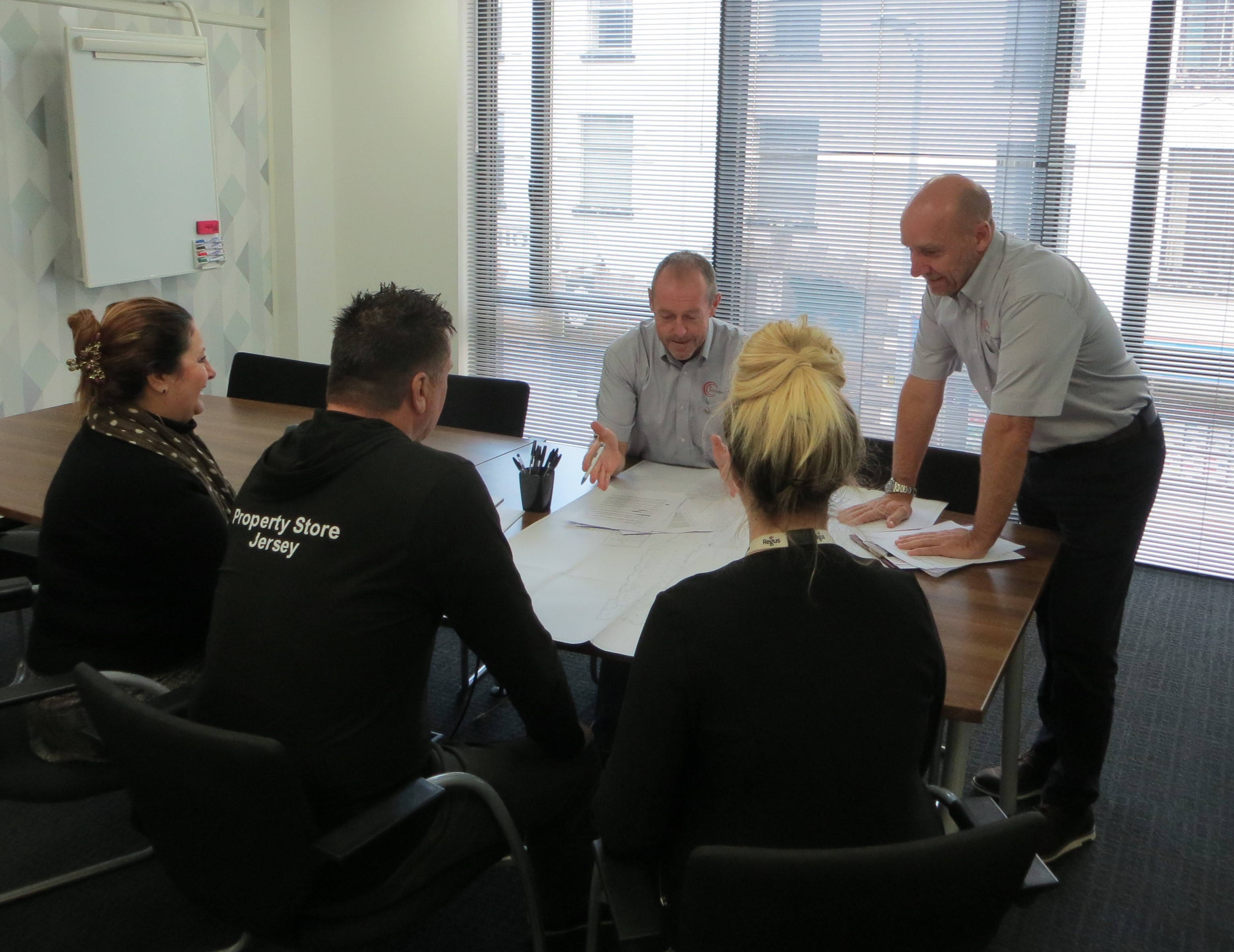 Five people in a meeting room discussing documents spread on a table near a window with blinds.
