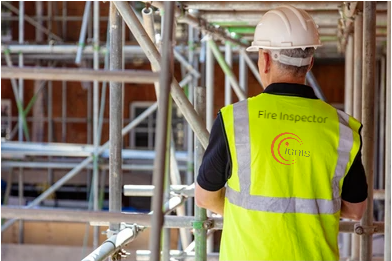 Fire inspector wearing a white hard hat and yellow safety vest standing among scaffolding at a construction site.