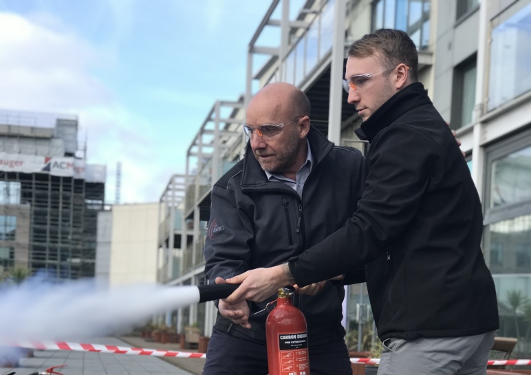 Two men wearing safety glasses operate a carbon dioxide fire extinguisher outside a modern building.