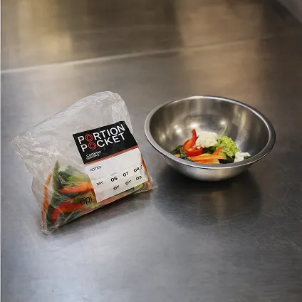 Clear plastic bag labeled Portion Pocket containing cut vegetables next to a metal bowl with sliced bell peppers and cauliflower on a stainless steel surface.