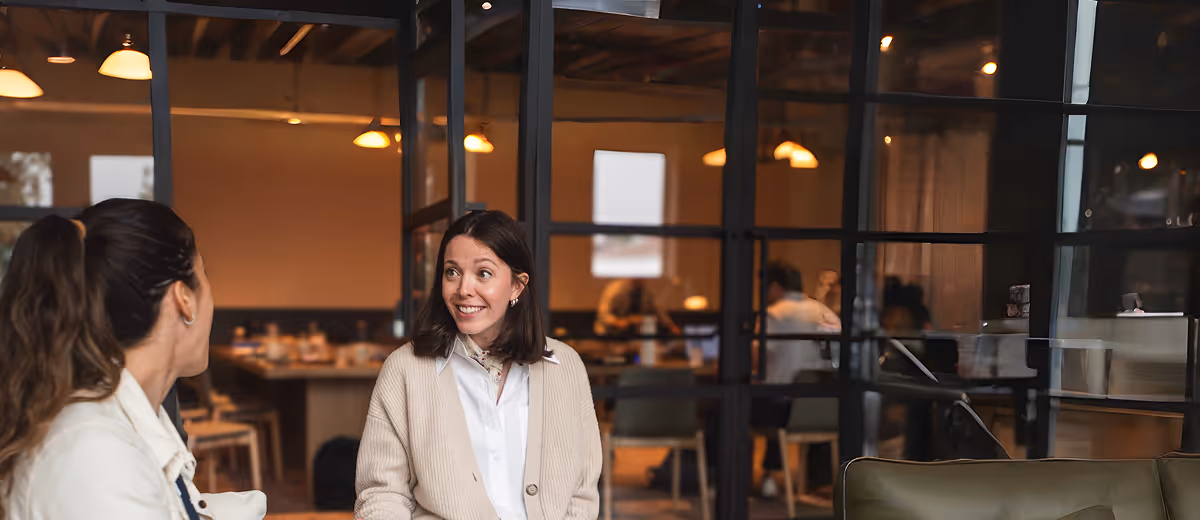 Two women having a friendly conversation in a modern office with glass walls and warm lighting.