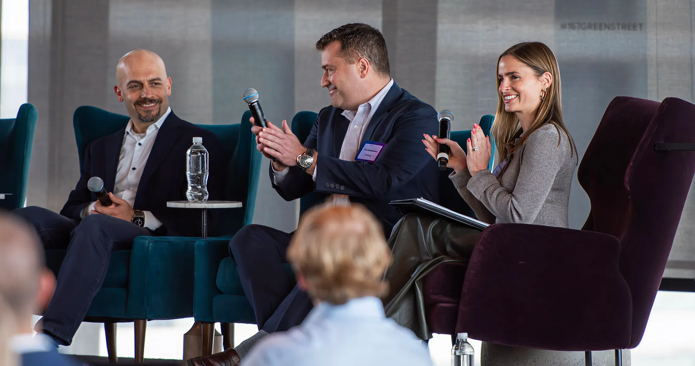 Three people seated on stage chairs holding microphones, smiling and clapping during a panel event.