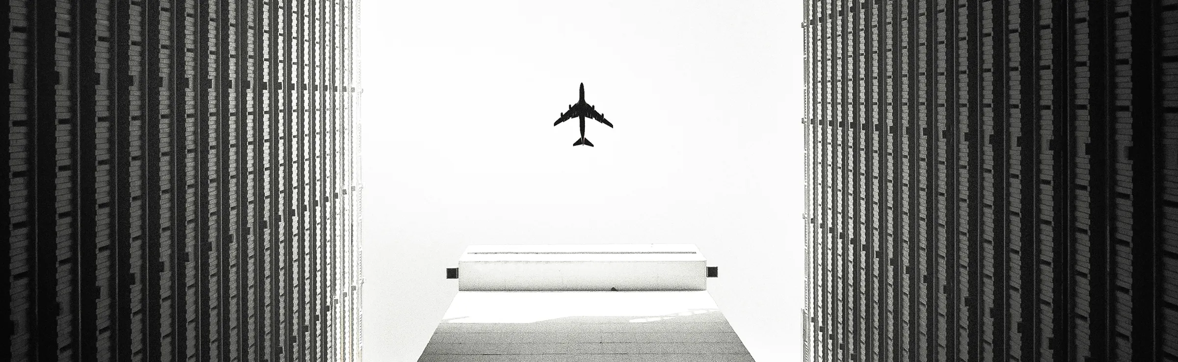 Silhouette of an airplane flying between two tall buildings against a bright sky.