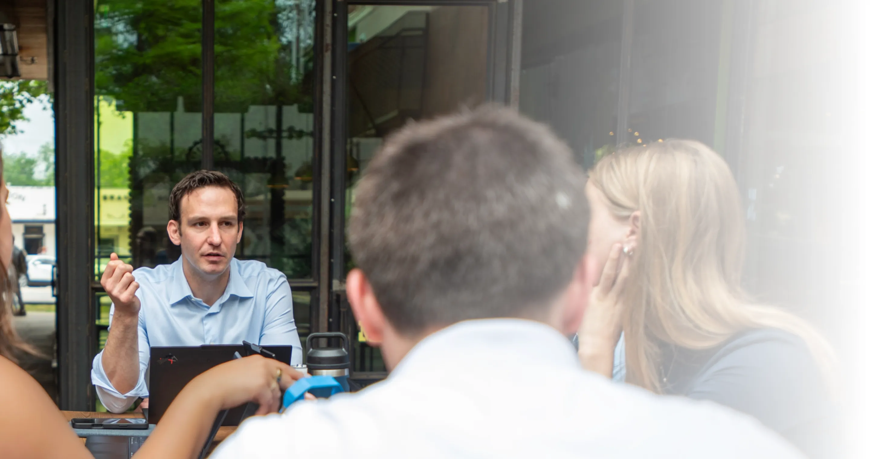 Man in blue shirt speaking to a group seated around a table in a bright indoor space.