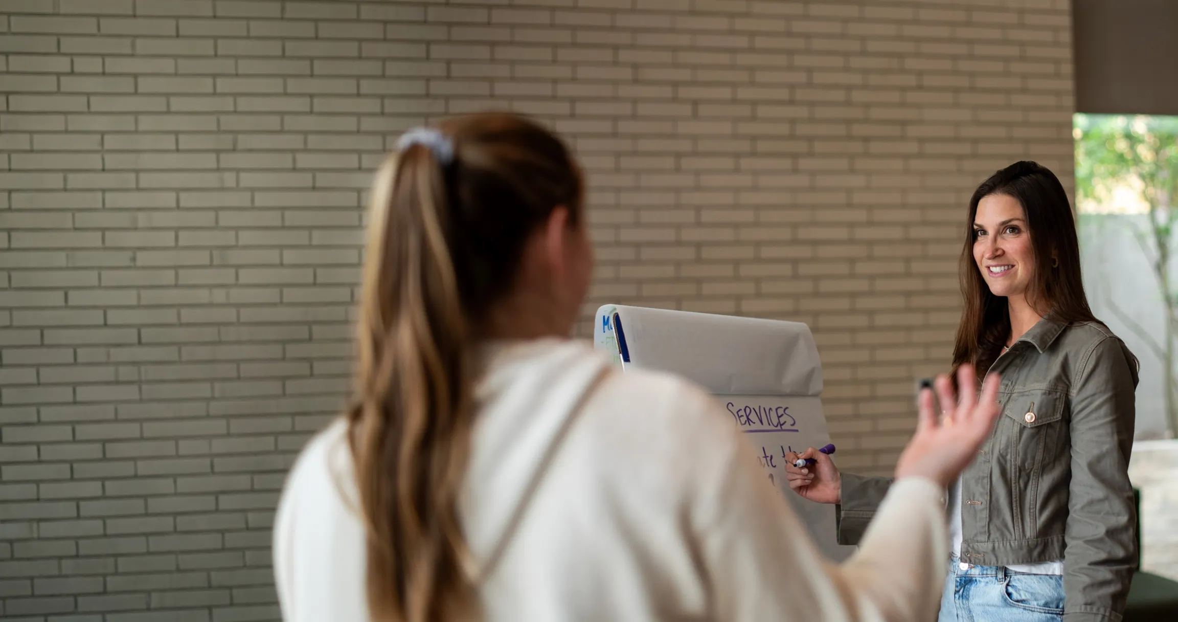 A woman writing on a flip chart while another woman gestures and talks to her indoors.