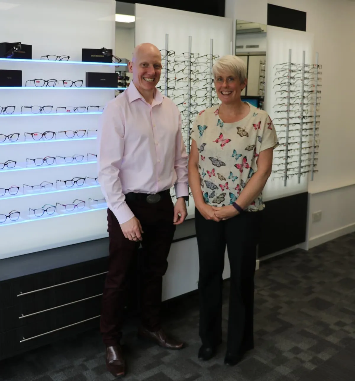 A man in a light pink shirt and dark pants standing next to a woman in a butterfly-patterned shirt and black pants in an eyewear store with glasses displayed on the walls.