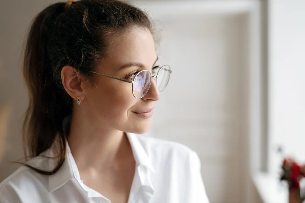 Young woman with ponytail wearing round glasses and a white shirt, looking out a window with a slight smile.
