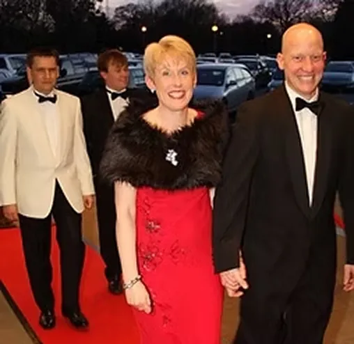 Four people dressed formally on a red carpet in the evening, two men in tuxedos, one woman in a red gown with a black fur stole, and one man in a white jacket.