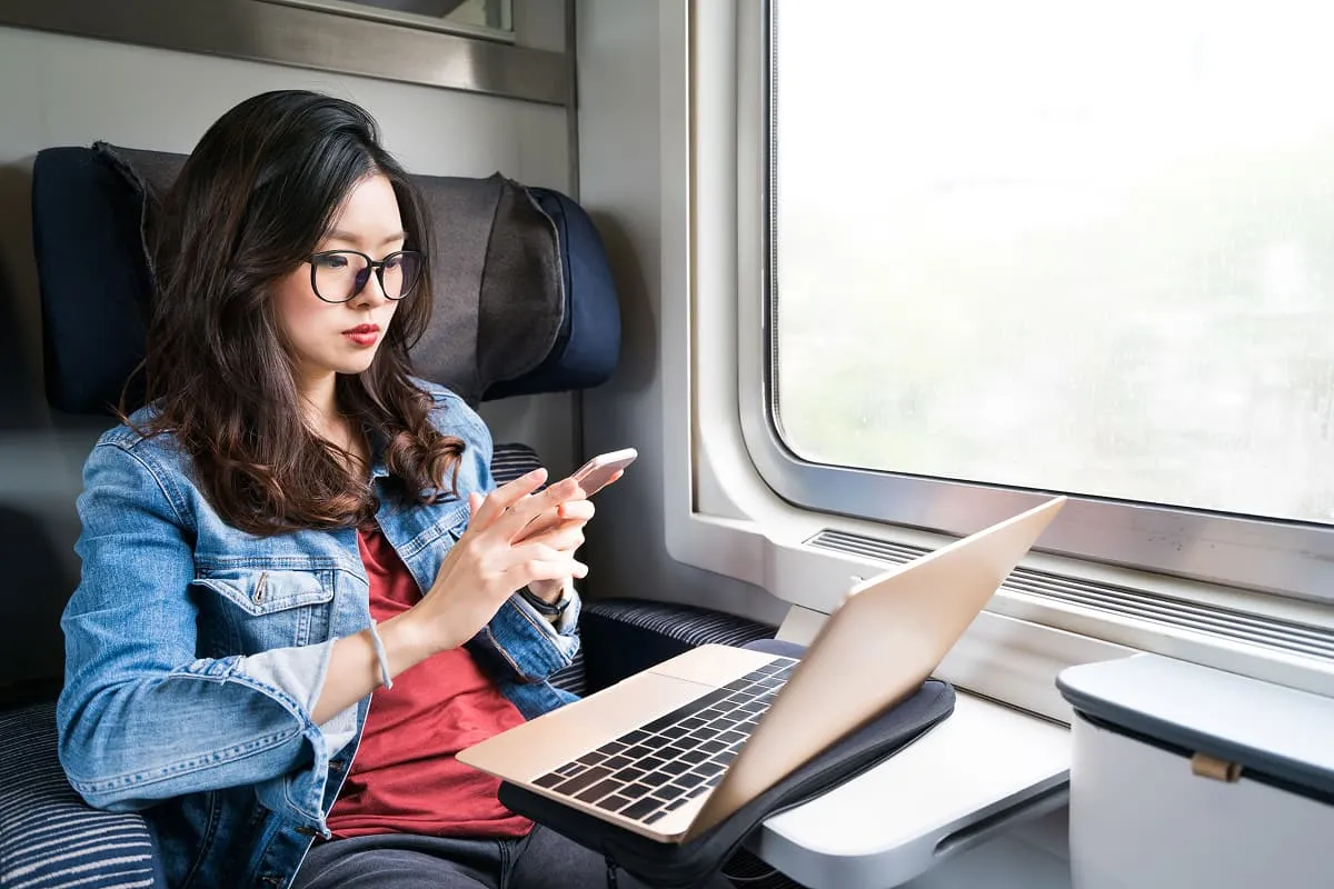 Woman wearing glasses and a denim jacket using her phone while sitting on a train with a laptop on the table by the window.