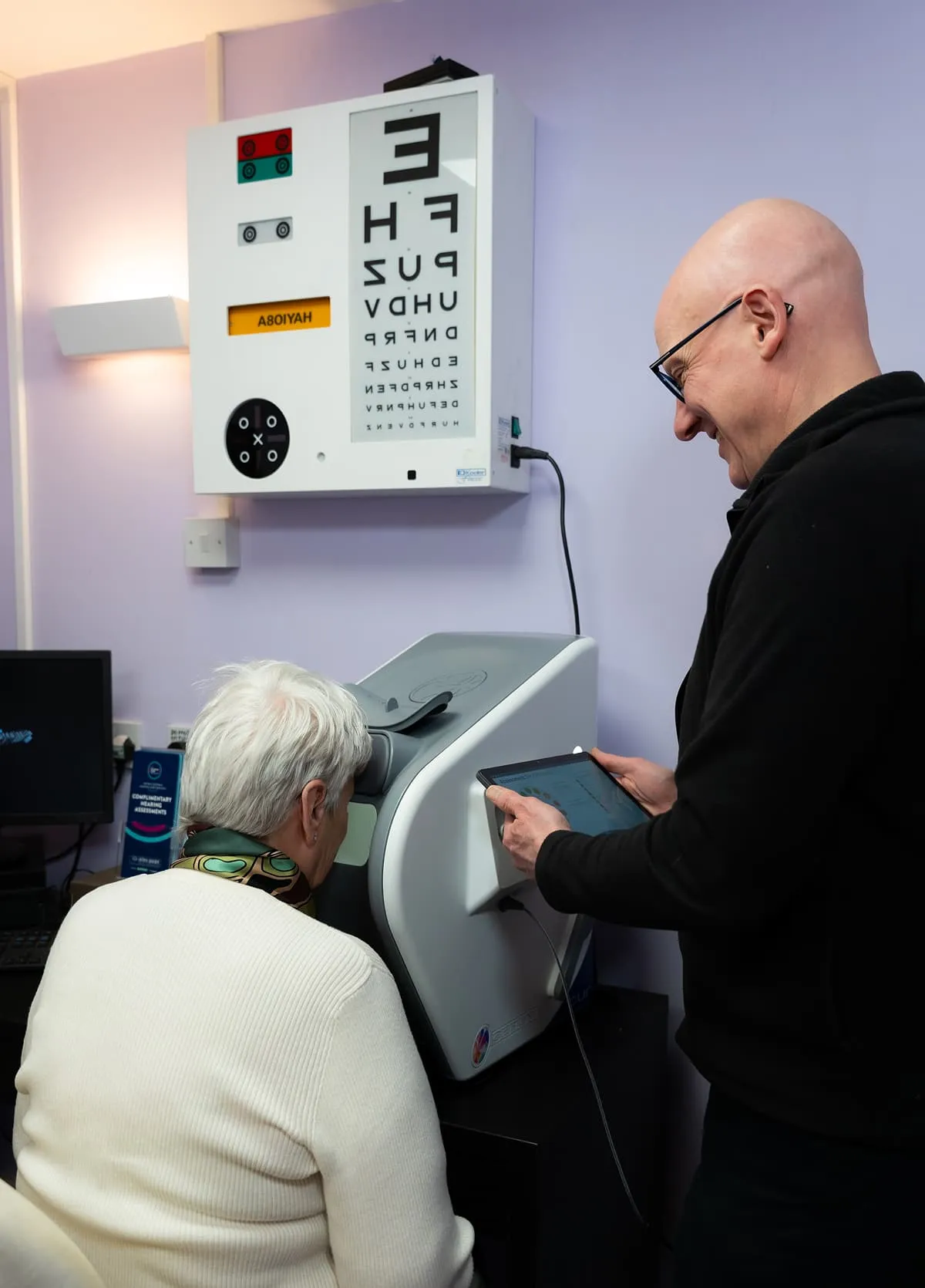 Older woman looking into eye testing machine while a man in glasses uses a tablet to assist her in an eye exam room.