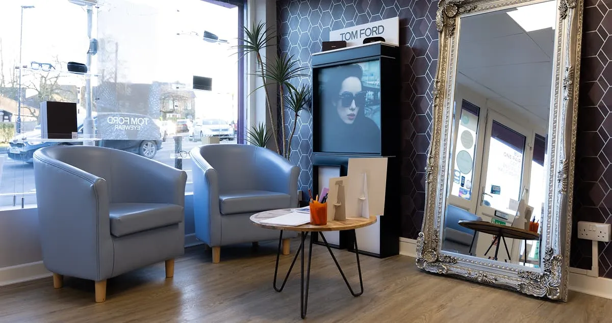 Modern waiting area with two light blue armchairs, a small round table with office supplies, a tall promotional display featuring a woman wearing sunglasses, and a large ornate silver-framed mirror.