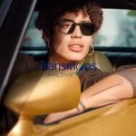 Person with curly hair wearing sunglasses and a brown top leaning on a yellow car's side mirror.