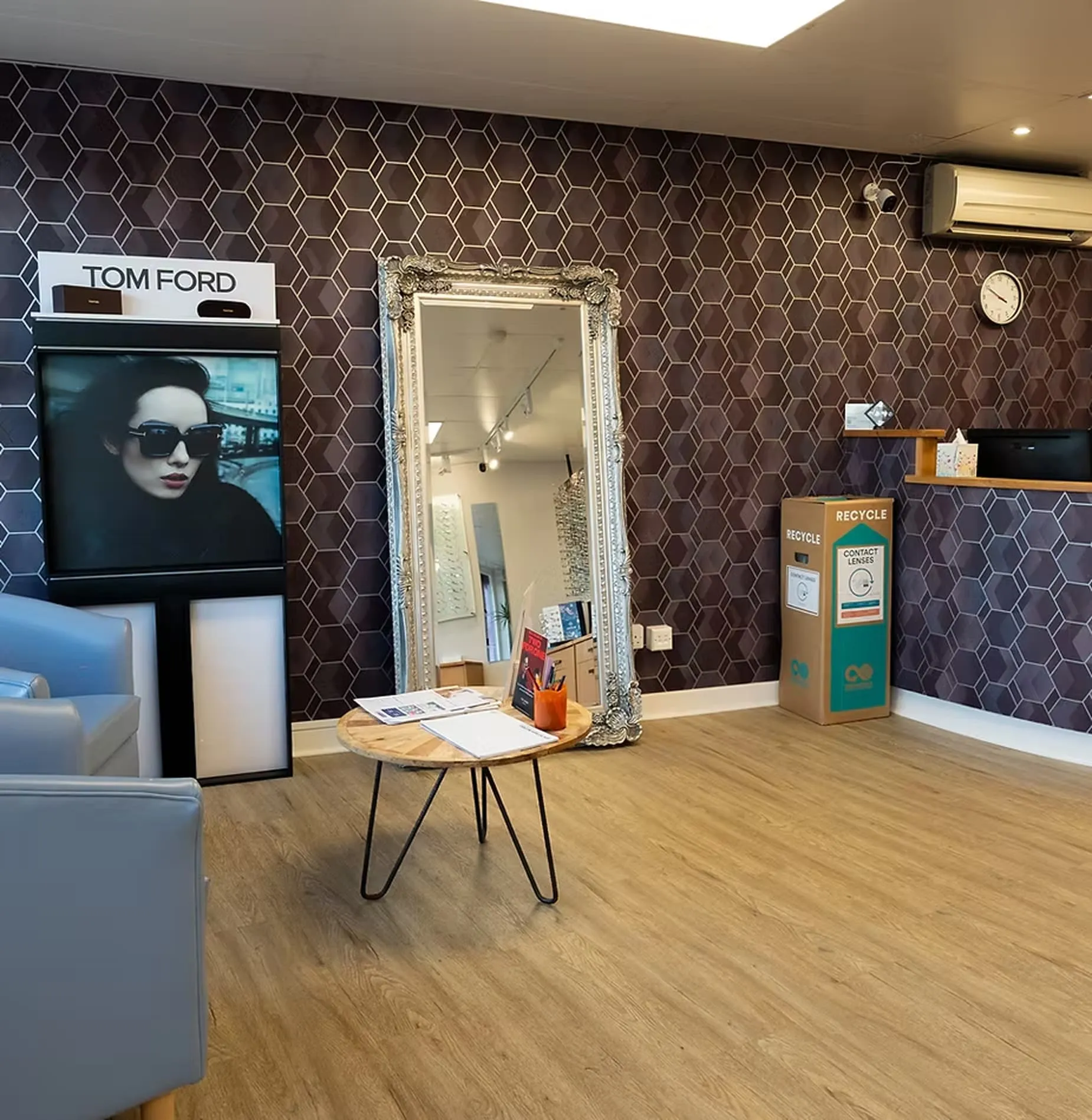 Sleek waiting area with gray chairs, a wooden round table, decorative mirror, recycle bin for contact lenses, and a Tom Ford advertisement on a patterned dark wall.