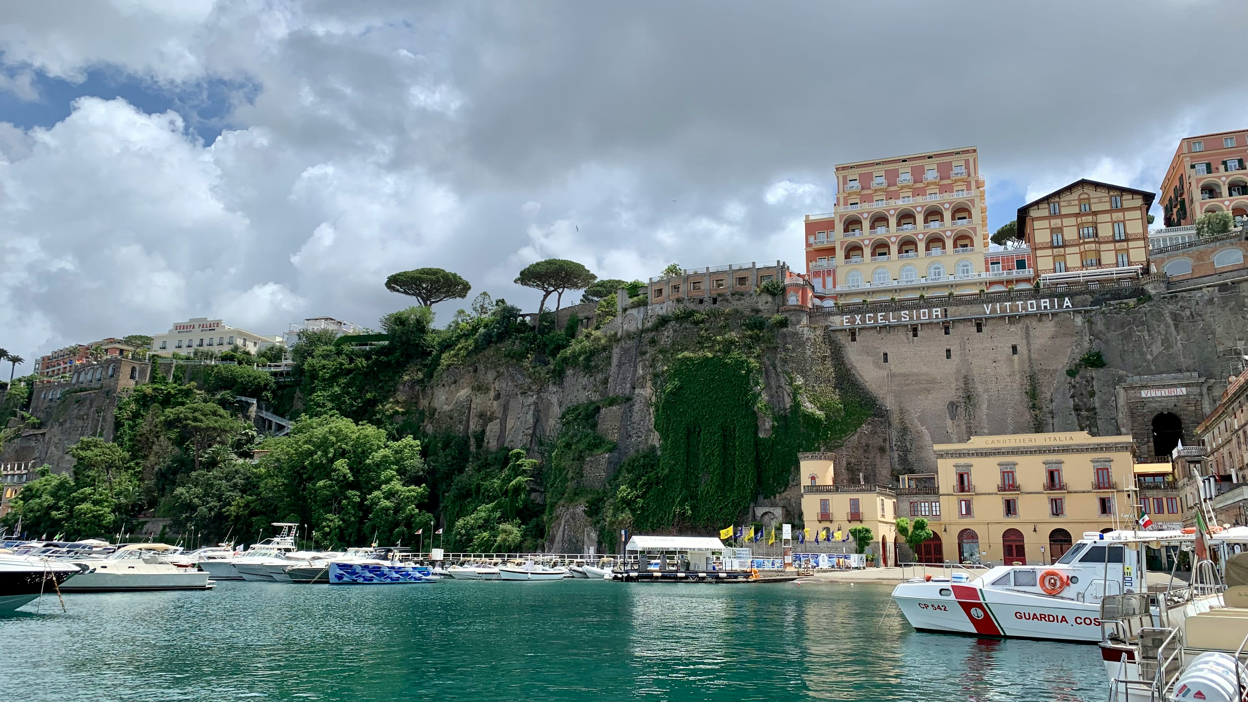Coastal scene with boats docked in turquoise water, lush green cliffs, and colorful buildings including one labeled 'Excelsior Vittoria'.