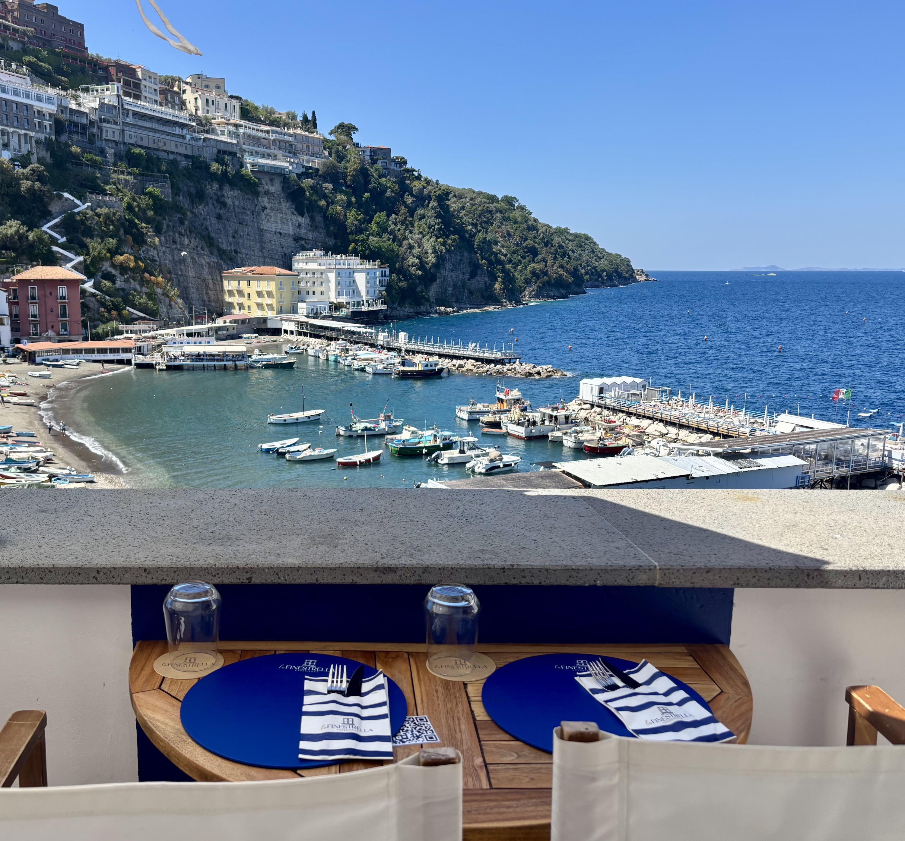Table set for two with napkins and glasses overlooking a sunny marina with boats and cliffside buildings.