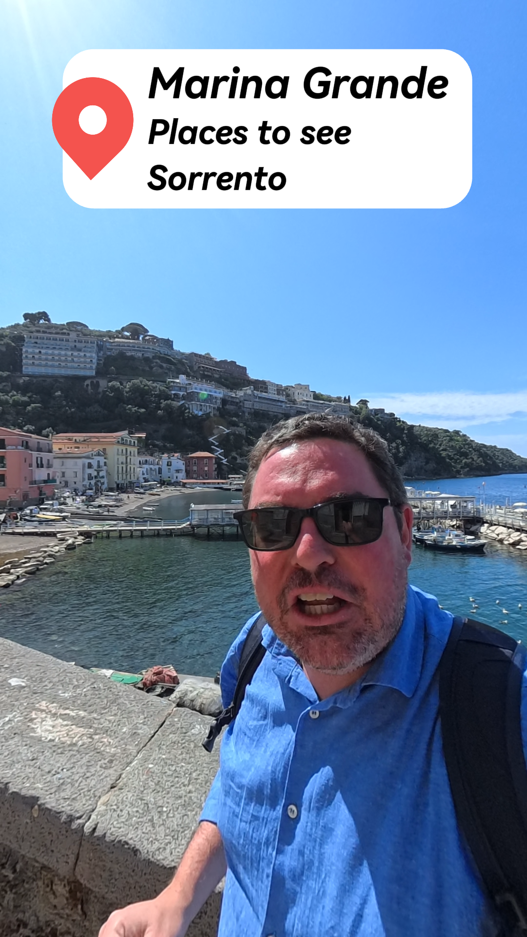 Man with sunglasses and a backpack standing by Marina Grande harbor in Sorrento on a sunny day.