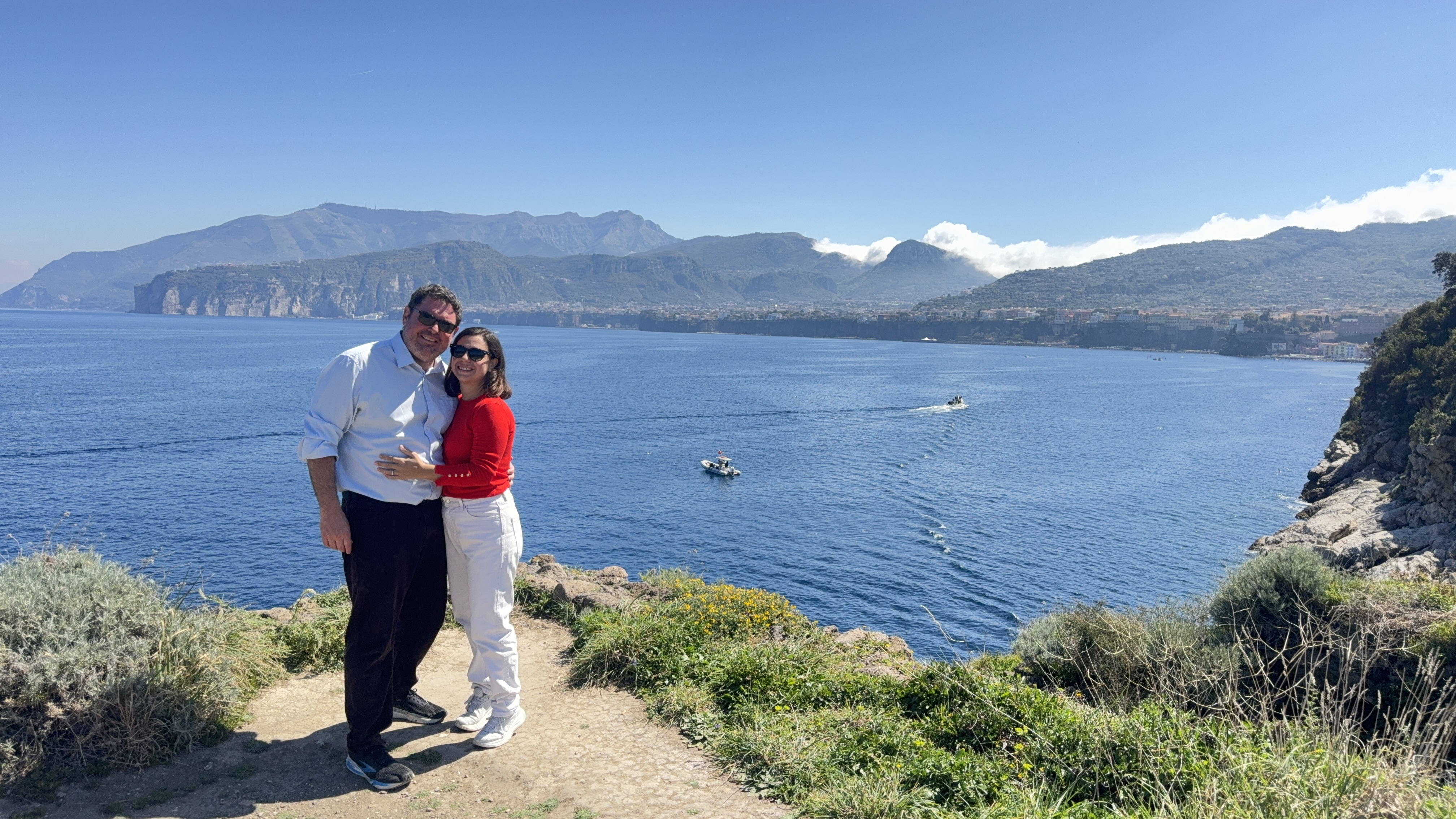 Couple embracing on a cliff overlooking a blue sea with boats and mountains in the background.