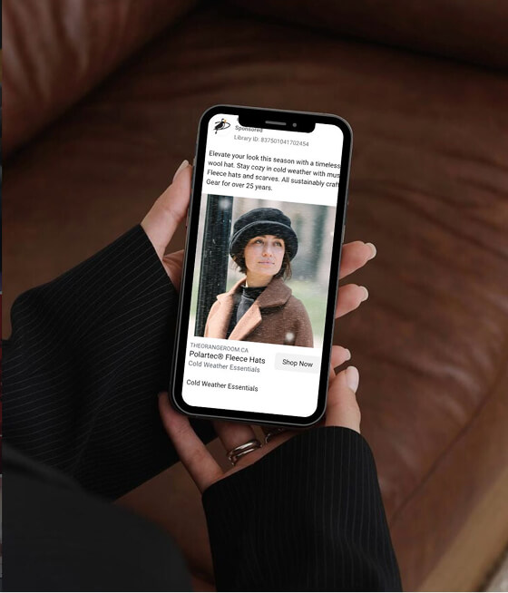 Hand holding a smartphone displaying an online ad for Polartec fleece hats with a woman wearing a black fleece hat and brown coat.
