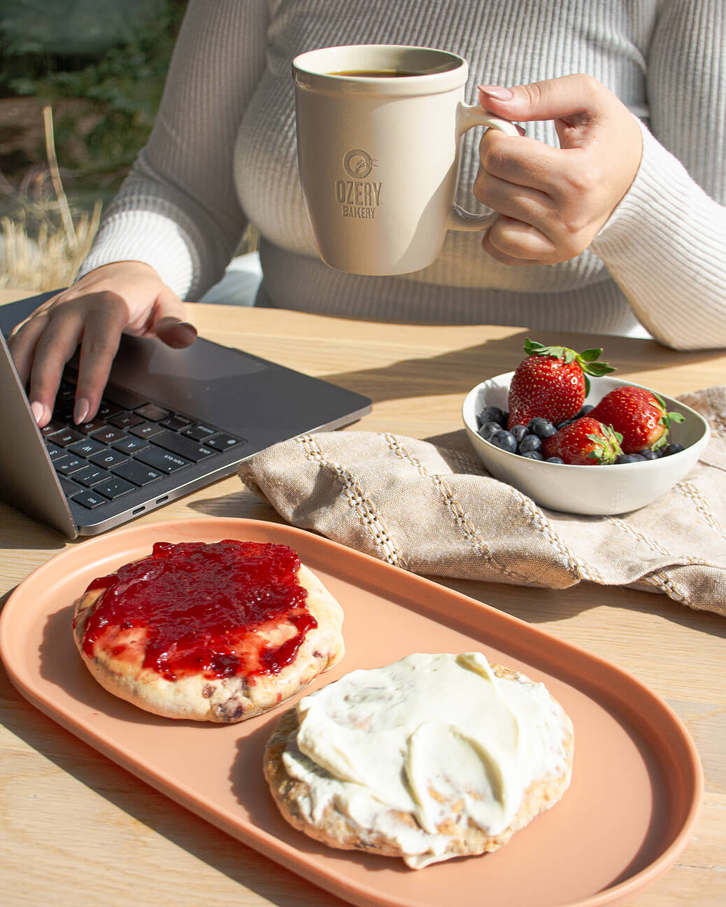 Person holding a beige Ozery Bakery mug in one hand and using a laptop with the other hand, with a peach plate of two bagel halves, one with cream cheese and the other with red jam, and a small bowl of strawberries and blueberries on the table.