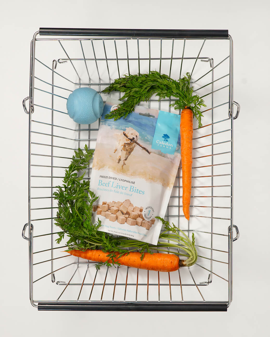 Metal basket containing a blue dog ball, a package of Caledon Farms freeze-dried beef liver bites with a dog image, and two fresh carrots with green tops.