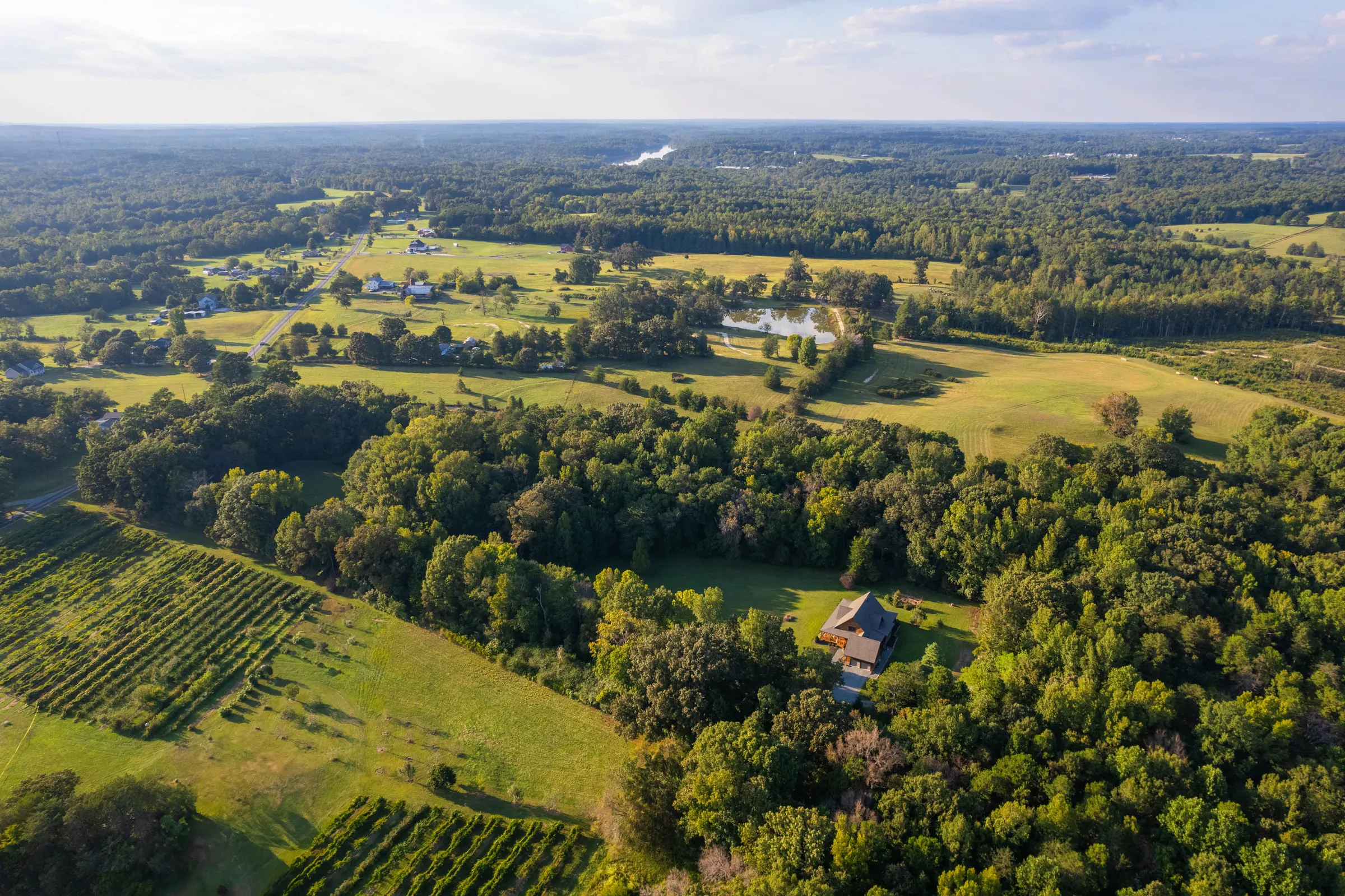 Aerial view of a Rolesville home amid open fields, mature trees, and a pond by Upper Echelon Visuals