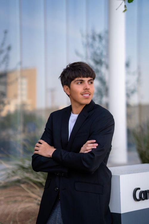 Young man in a black blazer with arms crossed looking to the side near a building with reflective glass.
