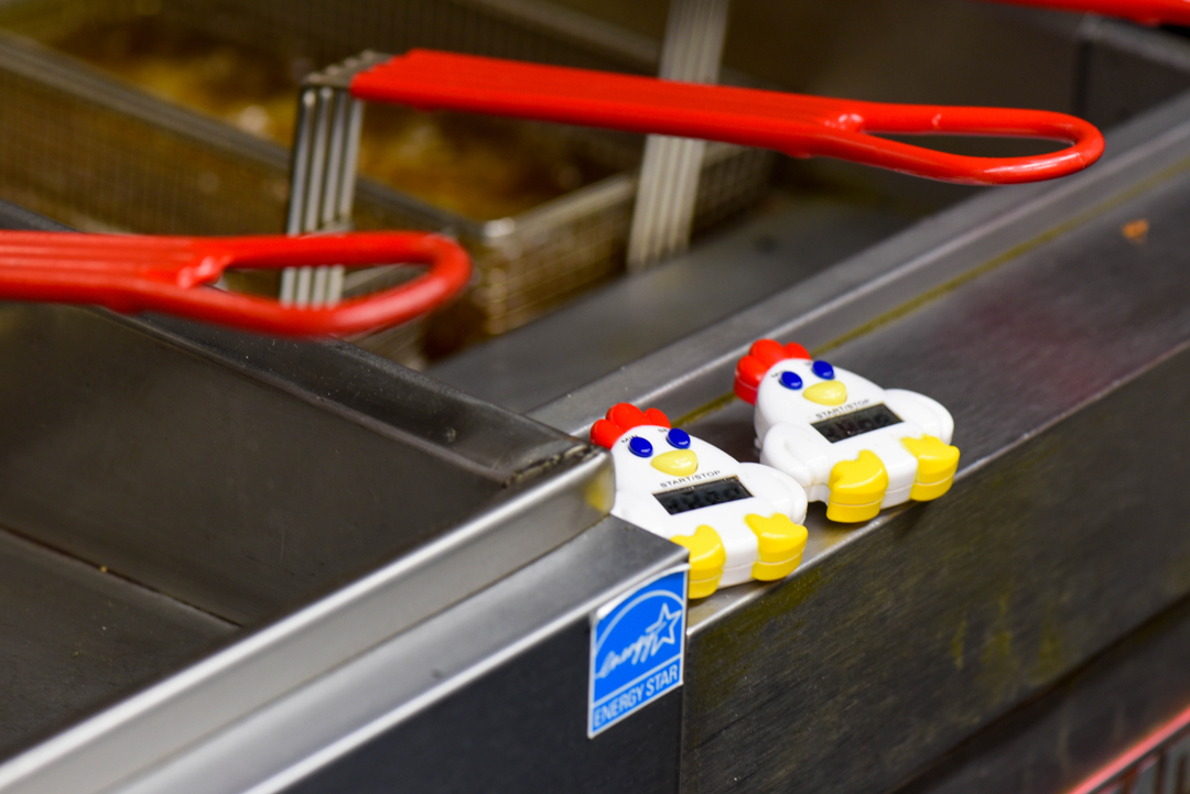 Two chicken-shaped kitchen timers placed on the edge of a commercial deep fryer with red handles visible in the background.