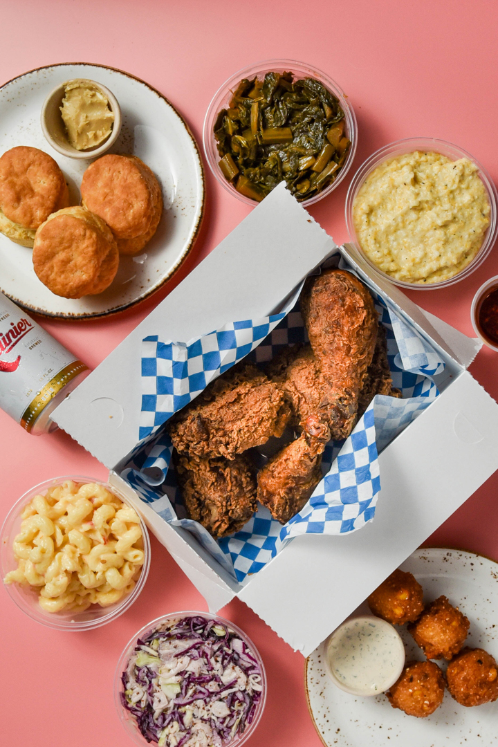 Box of fried chicken drumsticks surrounded by bowls of macaroni and cheese, collard greens, grits, coleslaw, biscuits with butter, and fried hush puppies with dipping sauce on a pink surface.