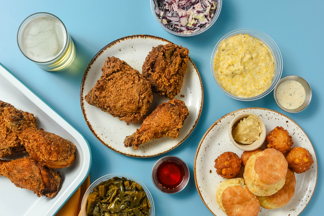 Table with plates of fried chicken, biscuits, hush puppies, collard greens, coleslaw, grits, dipping sauce, and a glass of beer on a blue surface.