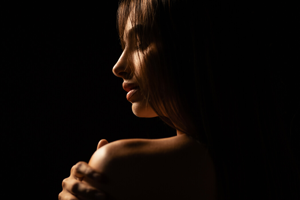Profile of a woman with soft lighting highlighting her face and shoulder against a dark background.