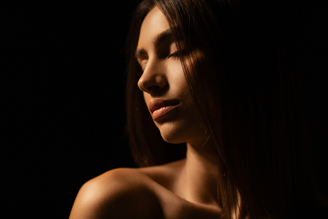 Close-up of a woman with closed eyes and long hair softly lit against a dark background.
