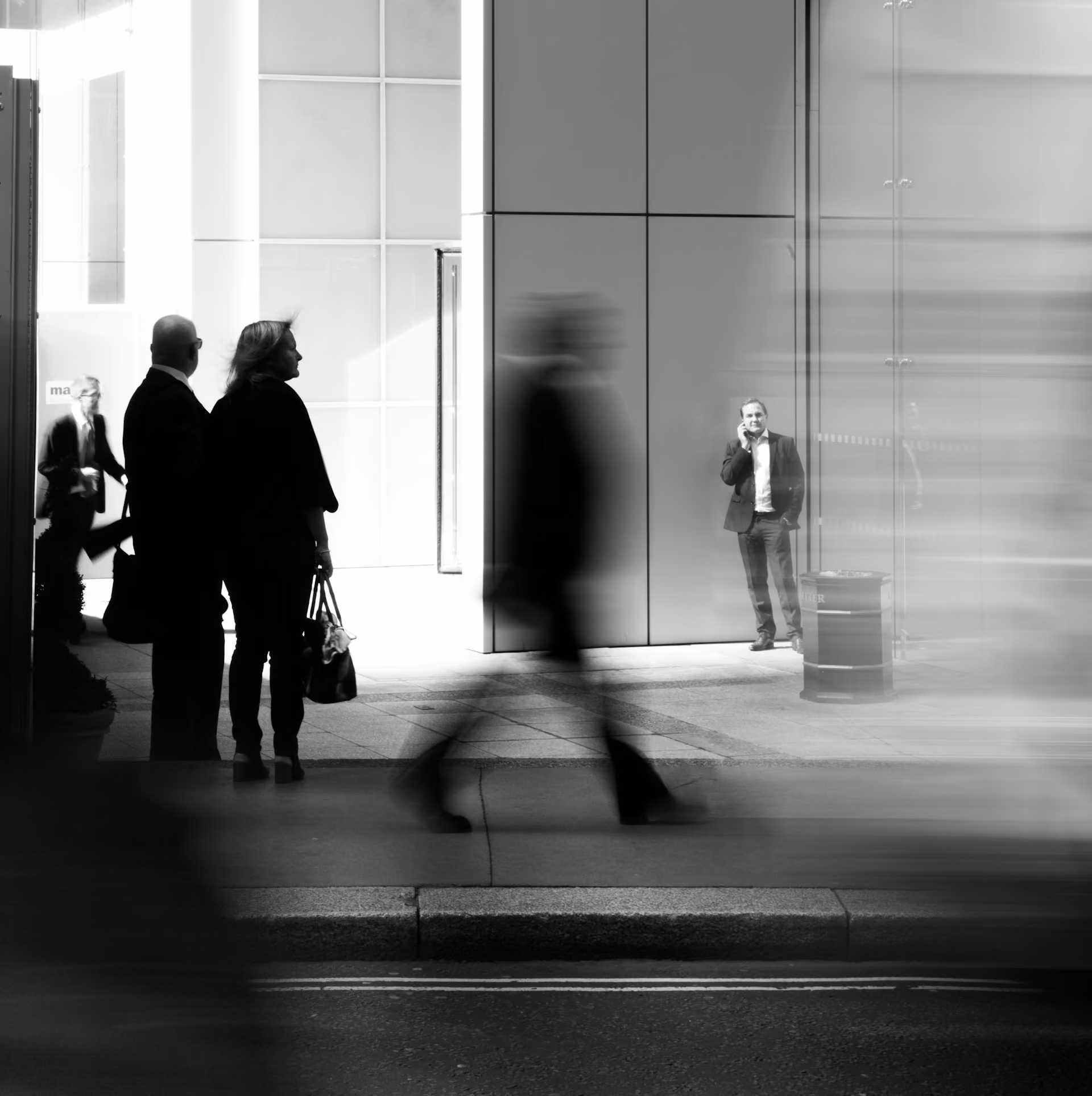 Black-and-white photo of business professionals outside a modern office building, with blurred figures in motion creating a sense of movement while a man stands in the background talking on the phone.