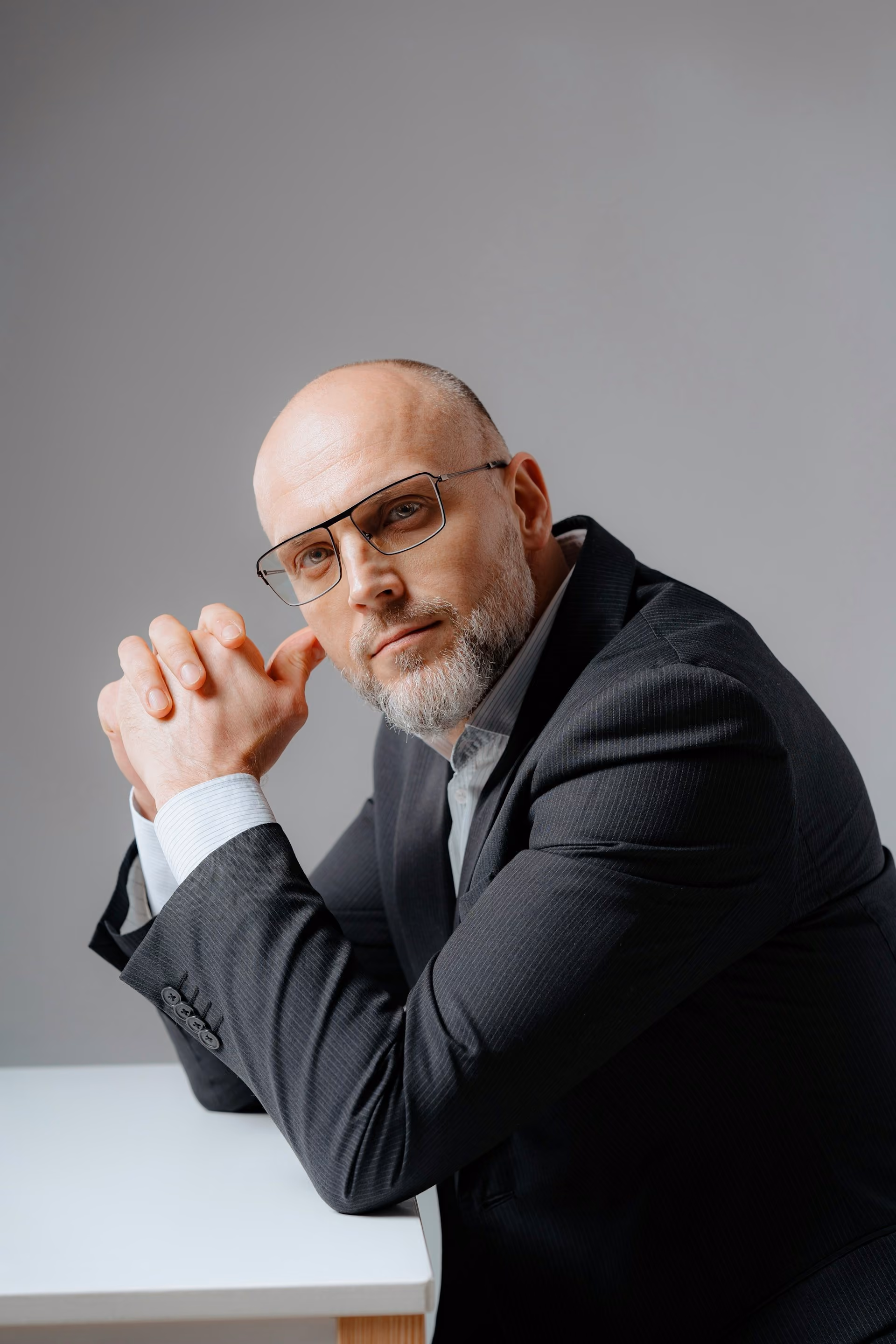 Business executive in a suit wearing glasses, seated with hands clasped and looking confidently at the camera.