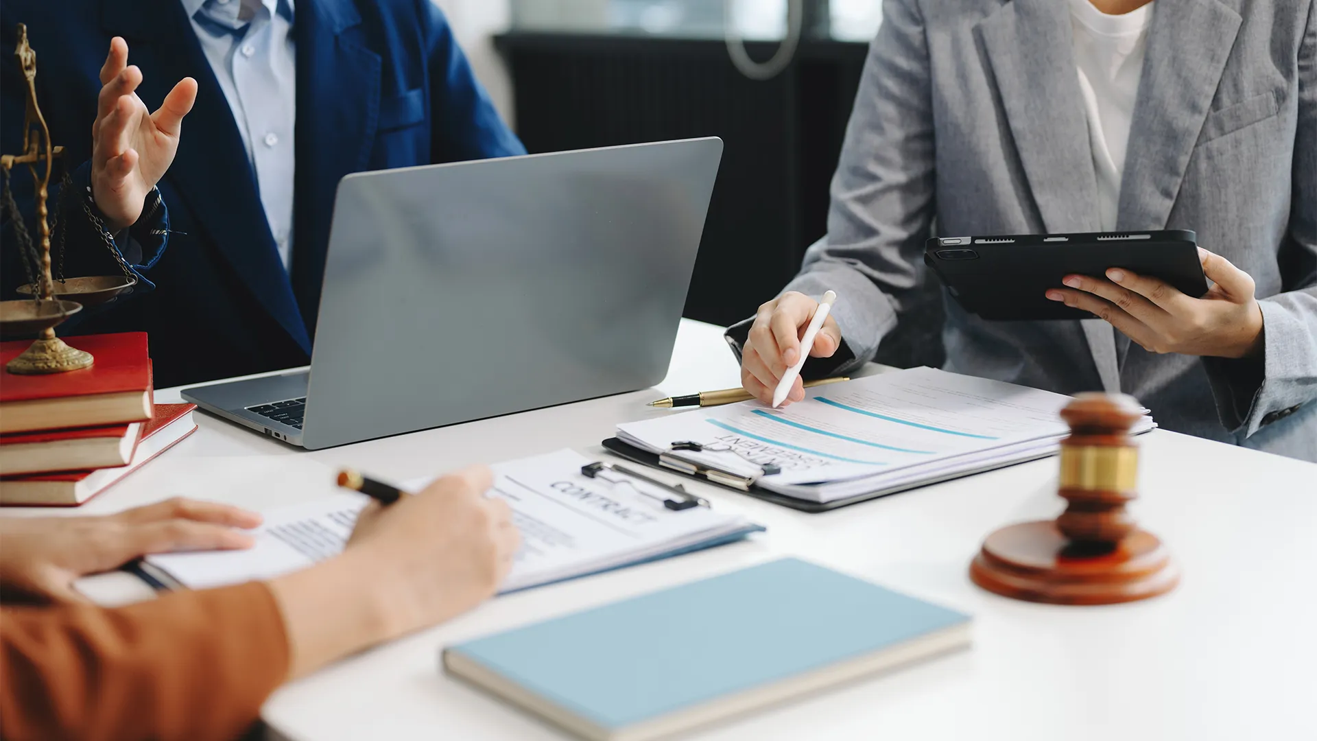 People in business attire discussing documents and using a laptop and tablet at a white office table, with legal books, a gavel, and a scale of justice visible.