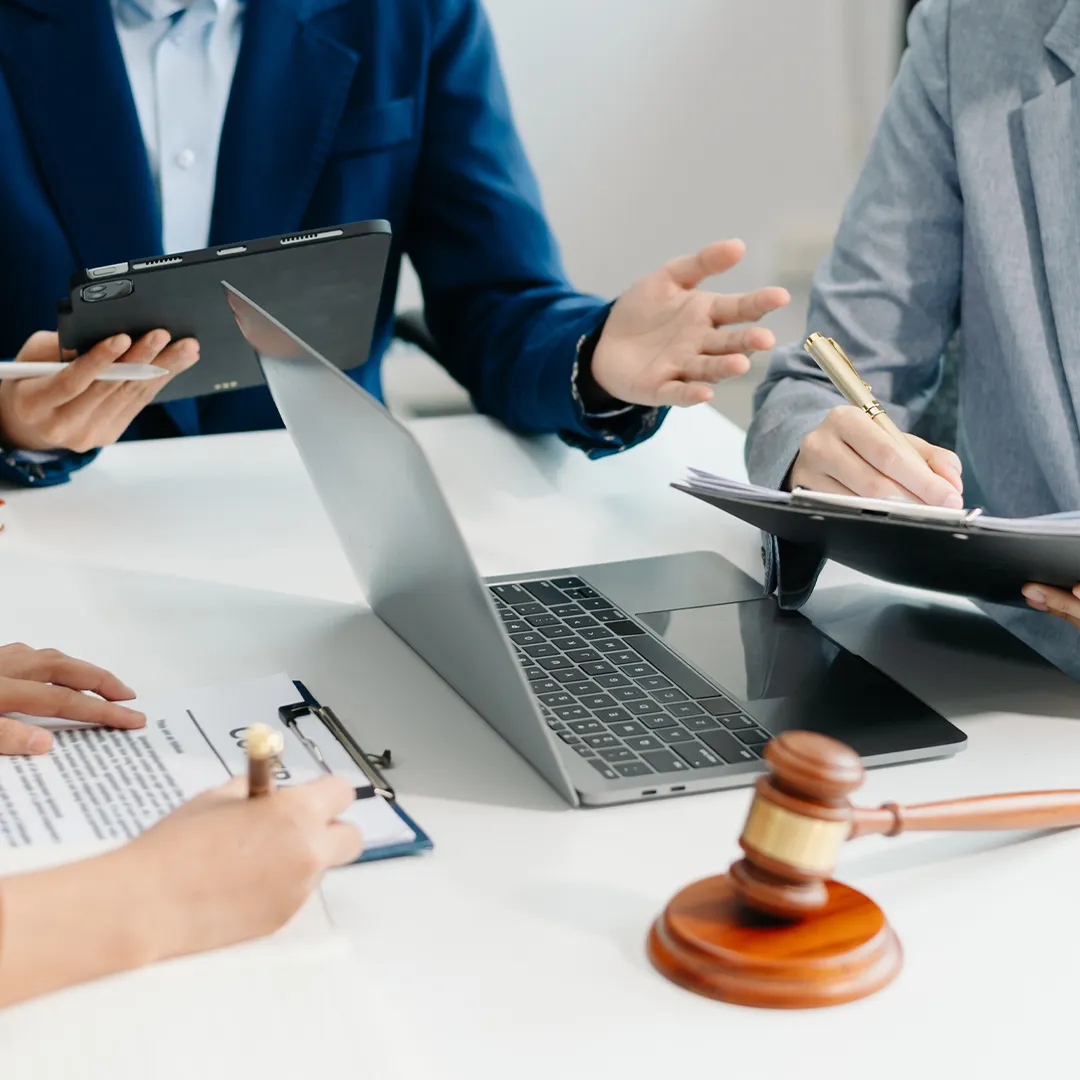 Three professionals in business attire discussing documents around a laptop on a white table with a wooden gavel nearby.