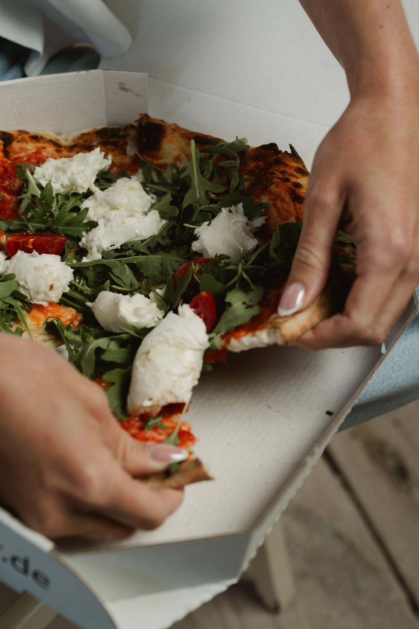 Hands holding an open pizza box topped with fresh vegetables and cheese.