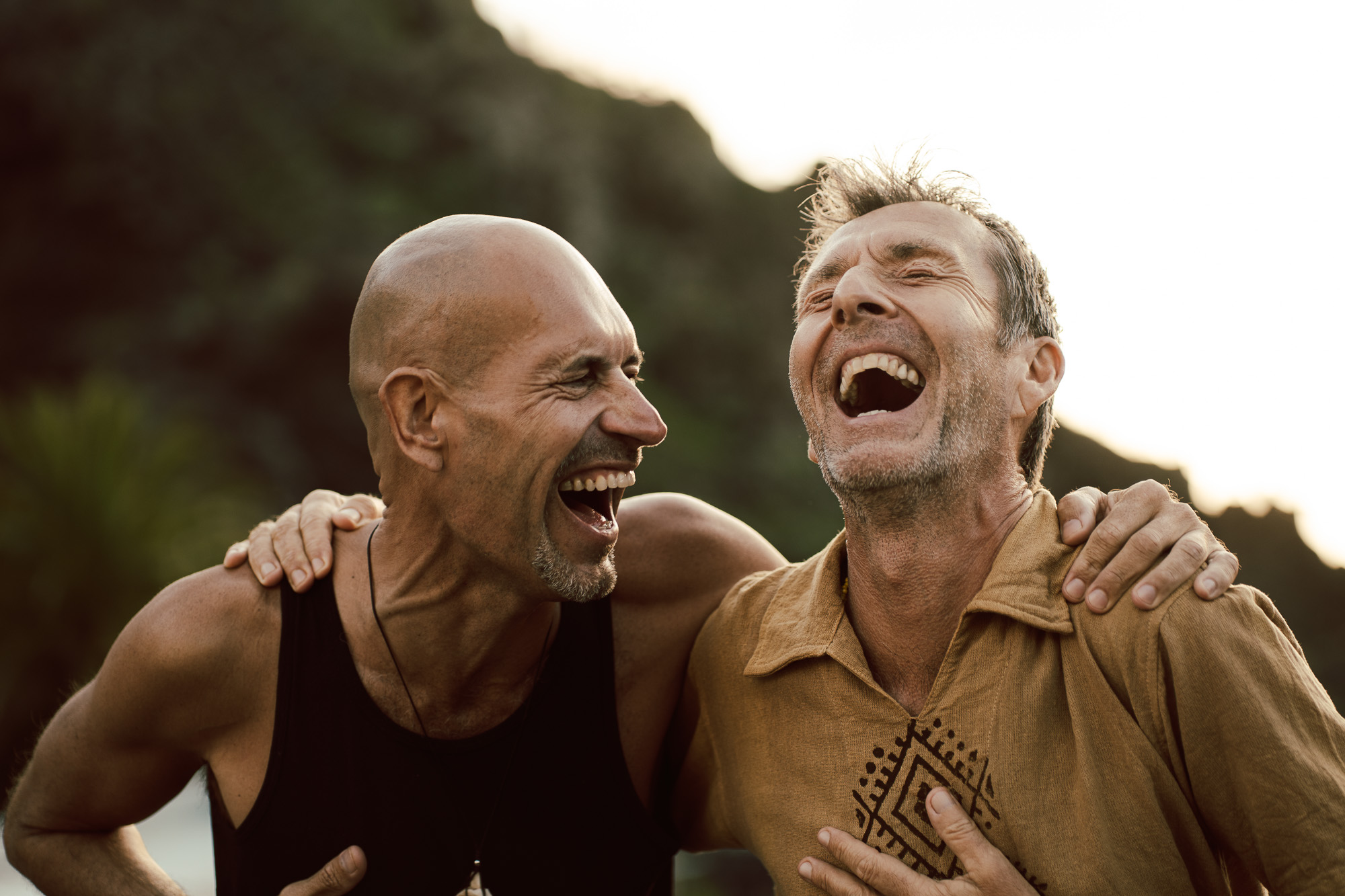 Two men laughing together with their arms around each other outdoors.