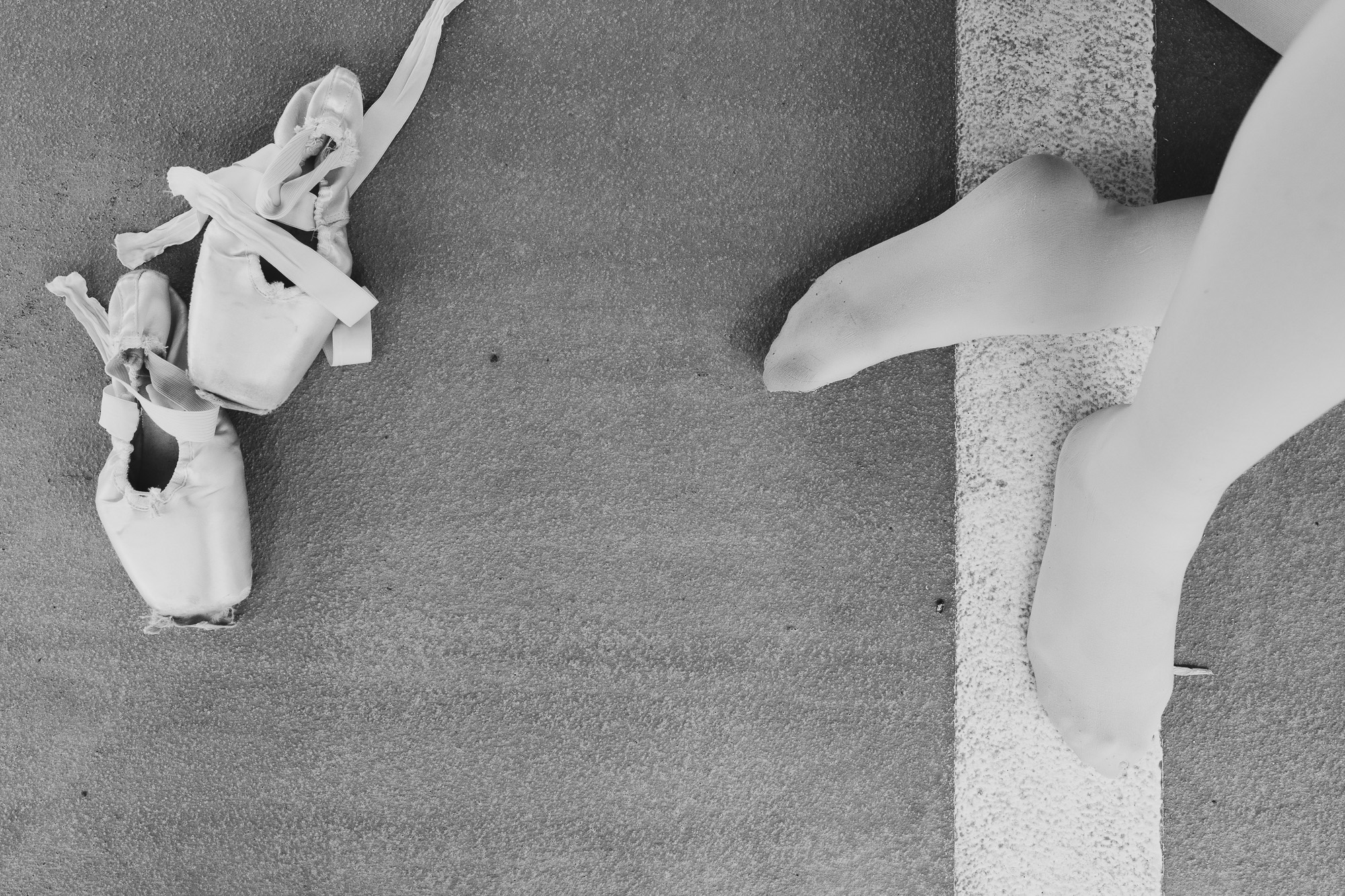 Black-and-white overhead view of ballet shoes and a dancer's legs on an industrial floor.