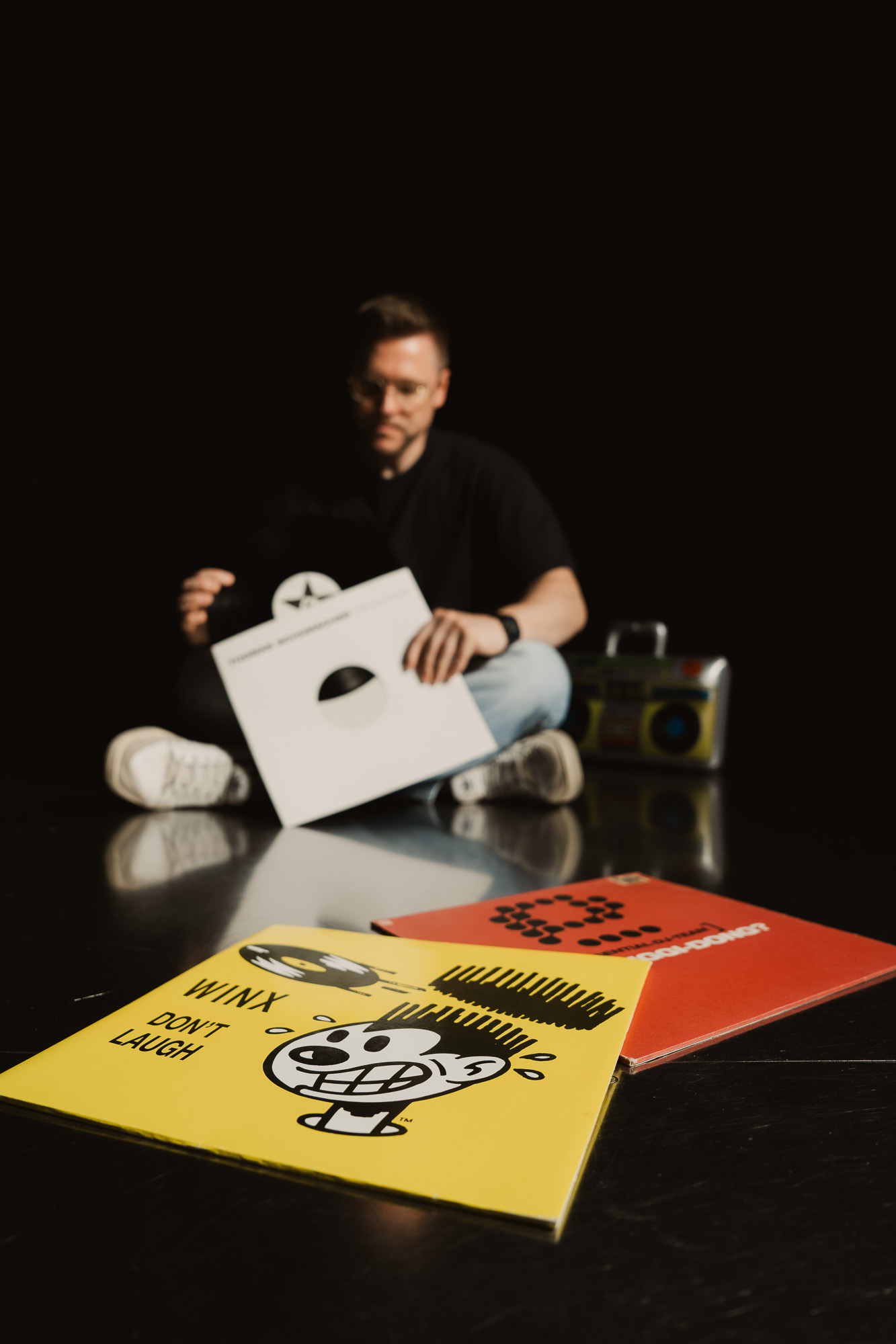 DJ surrounded by vinyl records and graphic album covers against a black background.