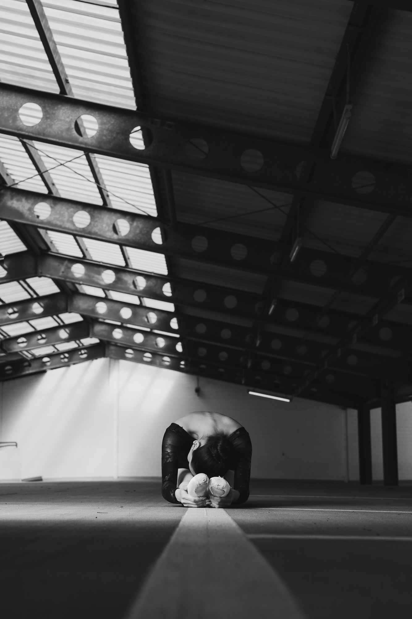 Black-and-white wide view of a lone dancer beneath the geometric roof of a vast industrial hall.