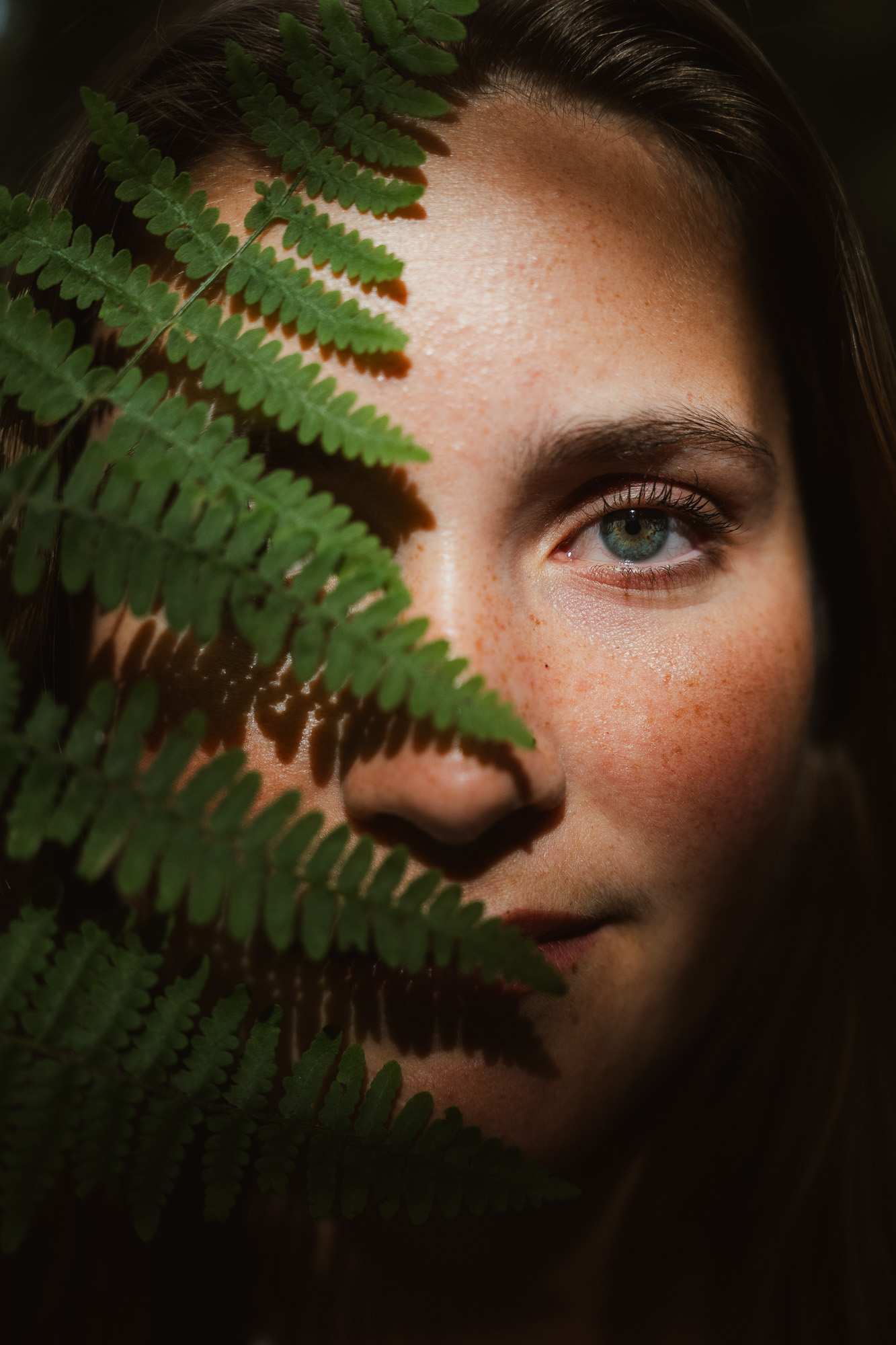 Close-up portrait of a woman partially hidden behind a green fern.