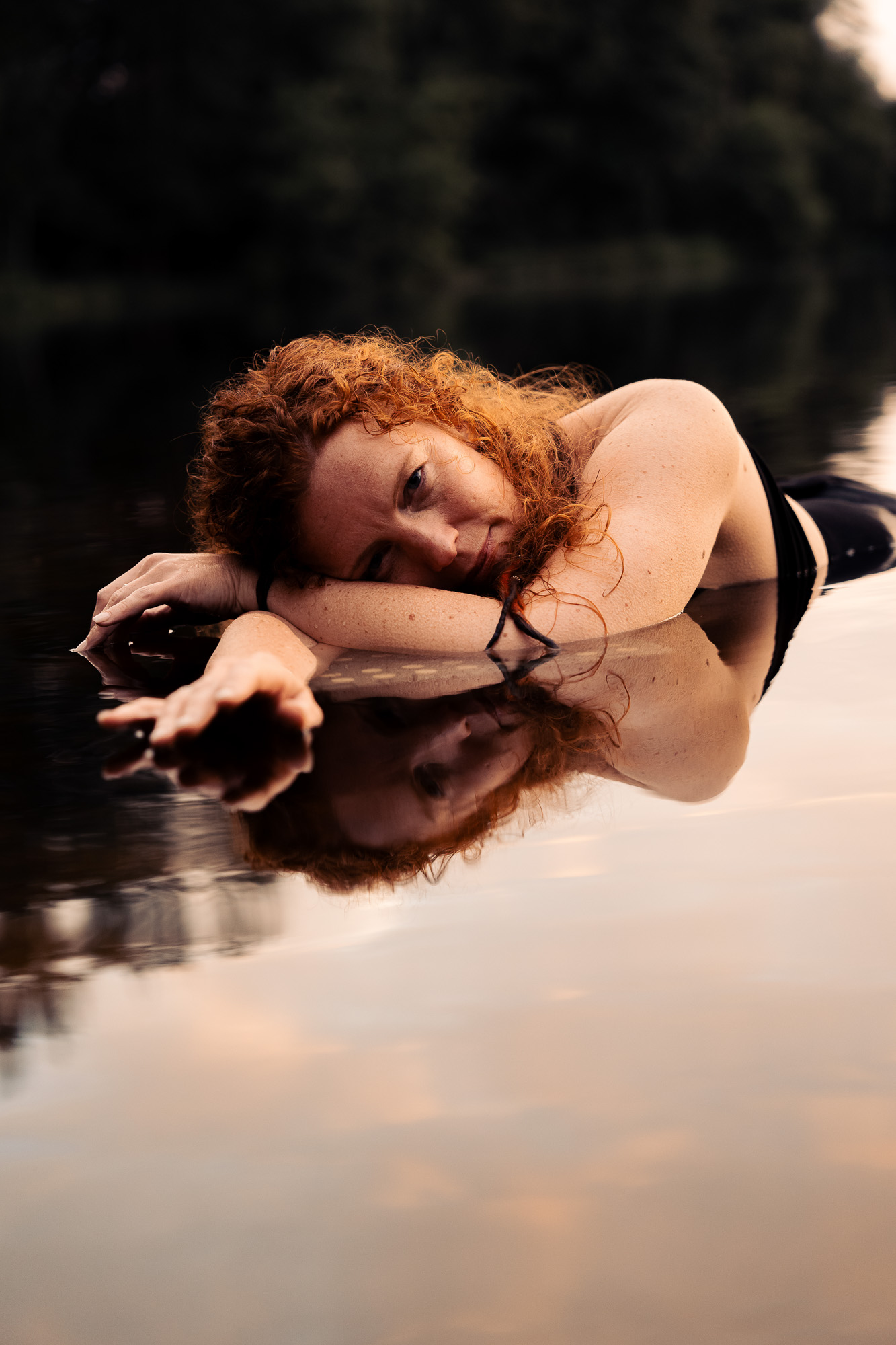 Red-haired woman lying beside a reflective water surface with her reflection beneath her.