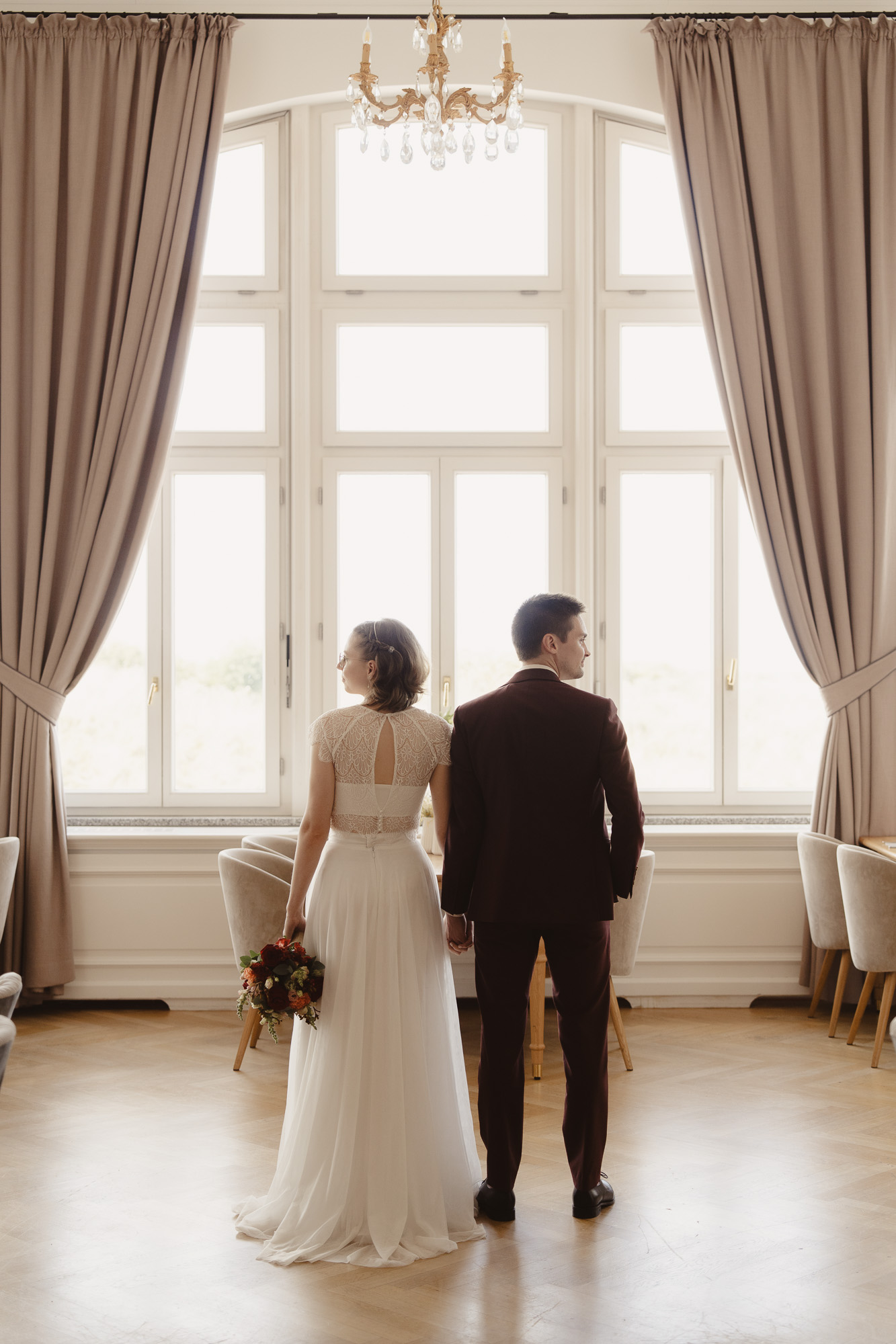 Bride and groom standing together in front of a tall window in an elegant room.