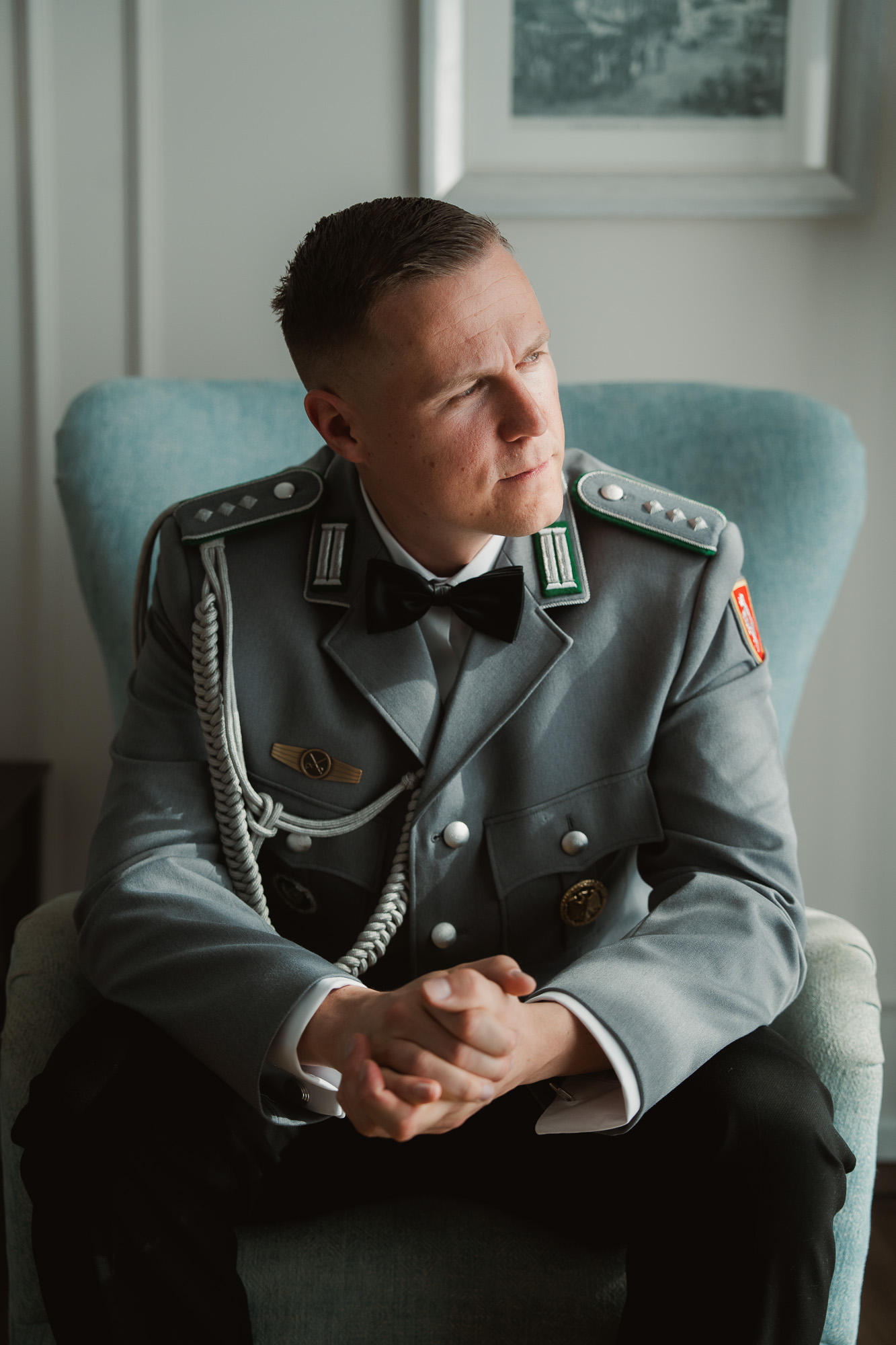 Groom in ceremonial uniform seated in an armchair beside a window.
