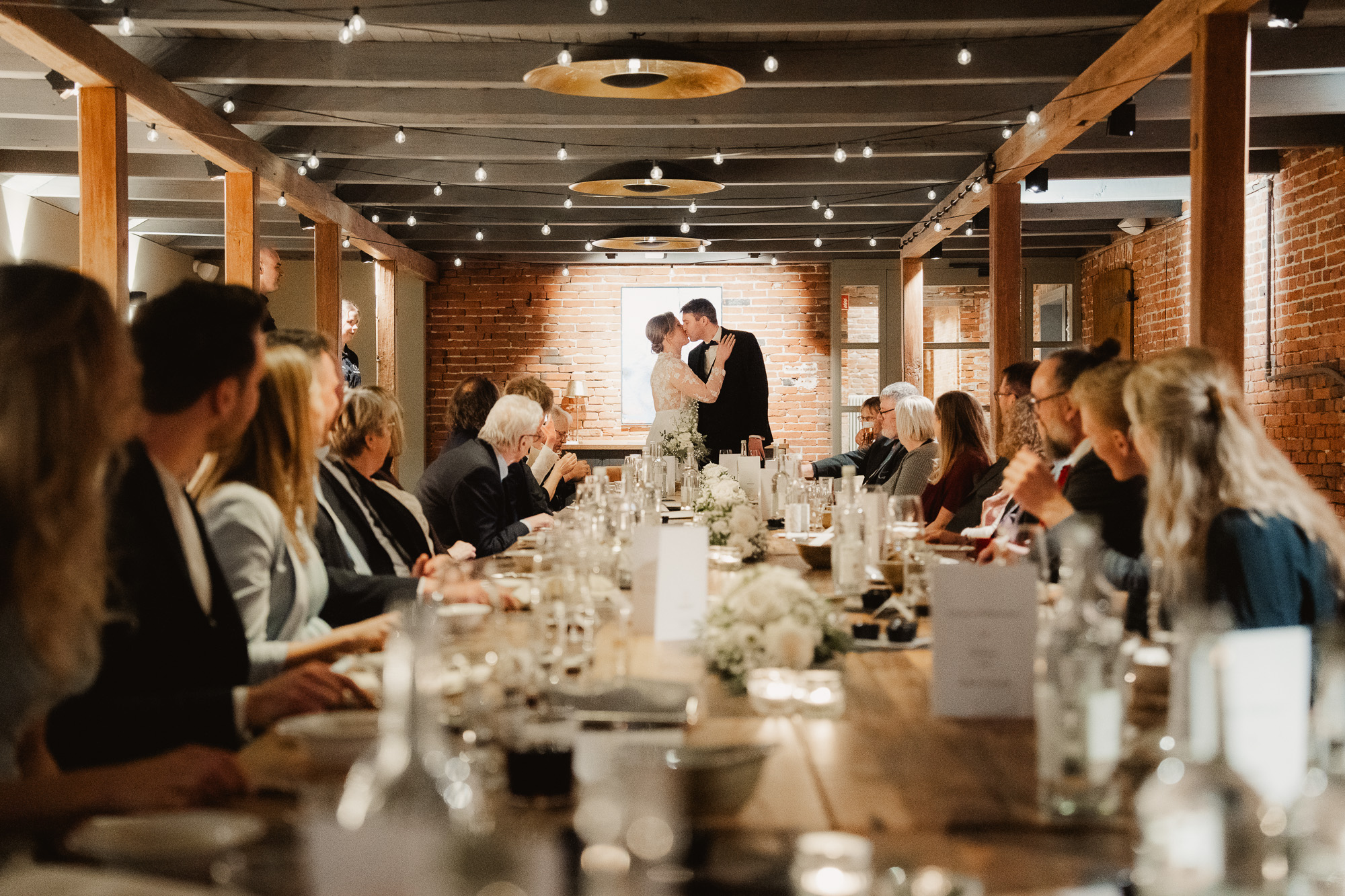 Long wedding dinner table filled with guests inside a warm modern reception venue.