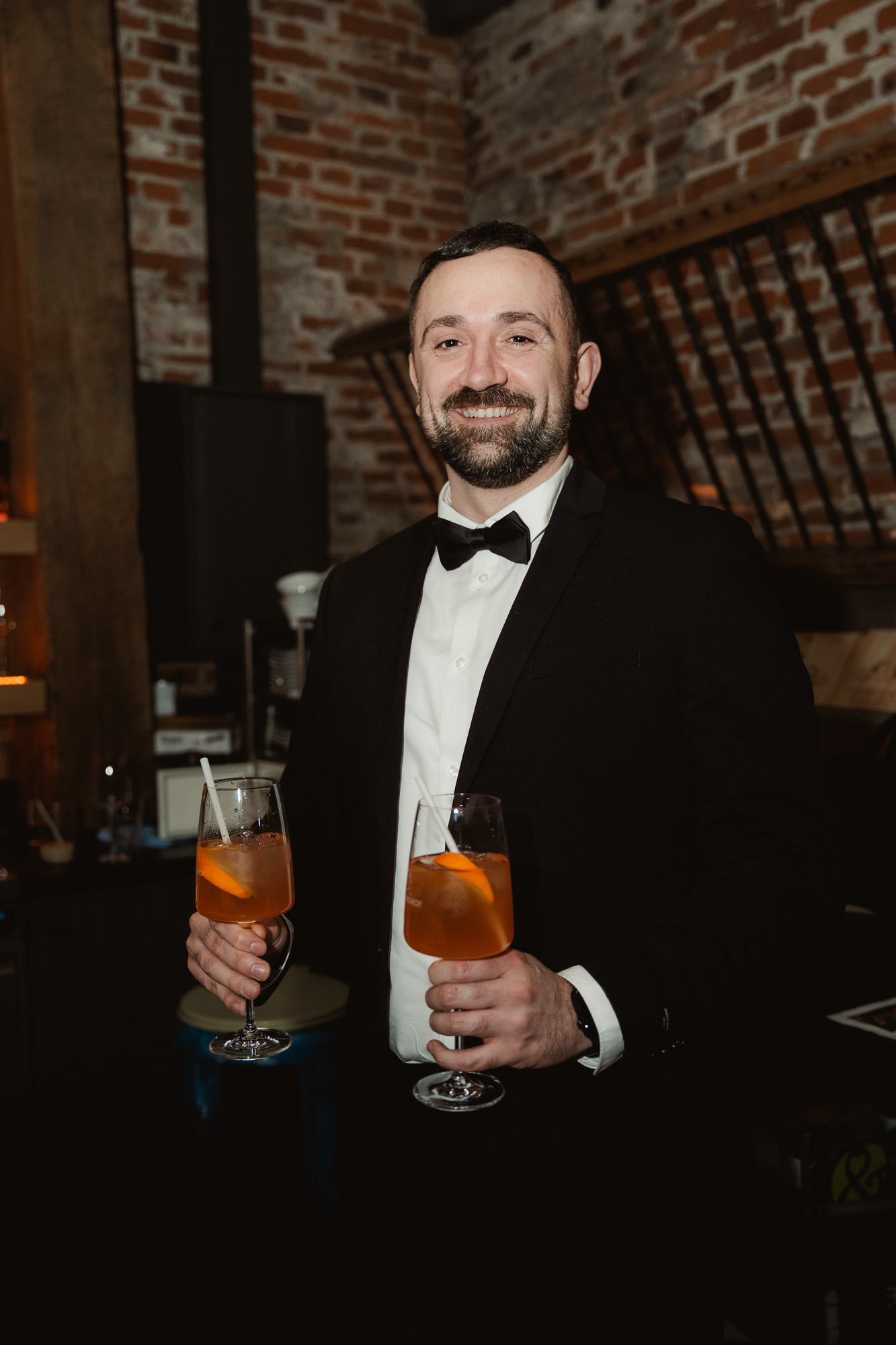 Groom in a black tuxedo smiling while holding two drinks at the wedding reception.