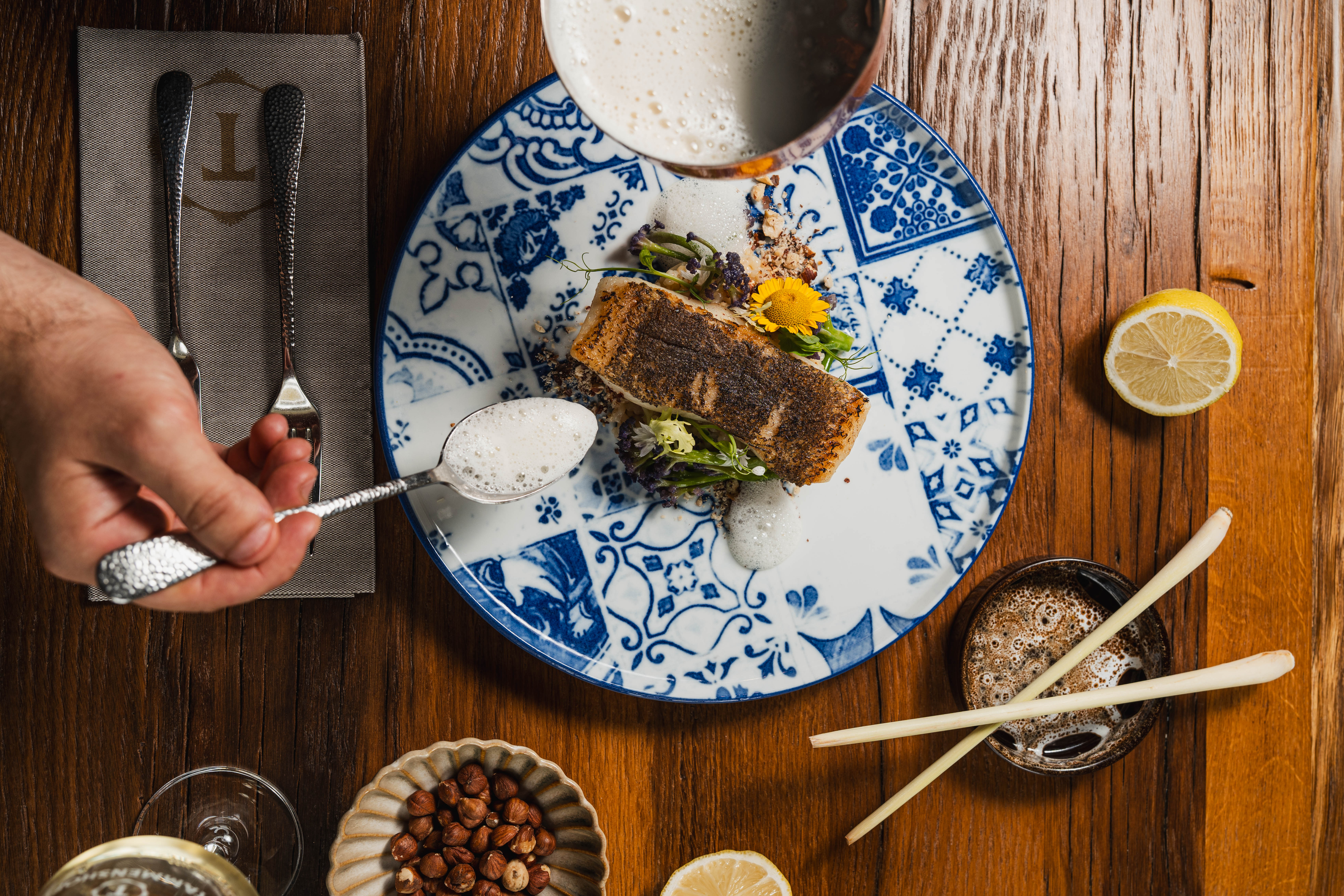 Overhead view of a fish dish with sauce, lemon, bread and side dishes on a wooden table.
