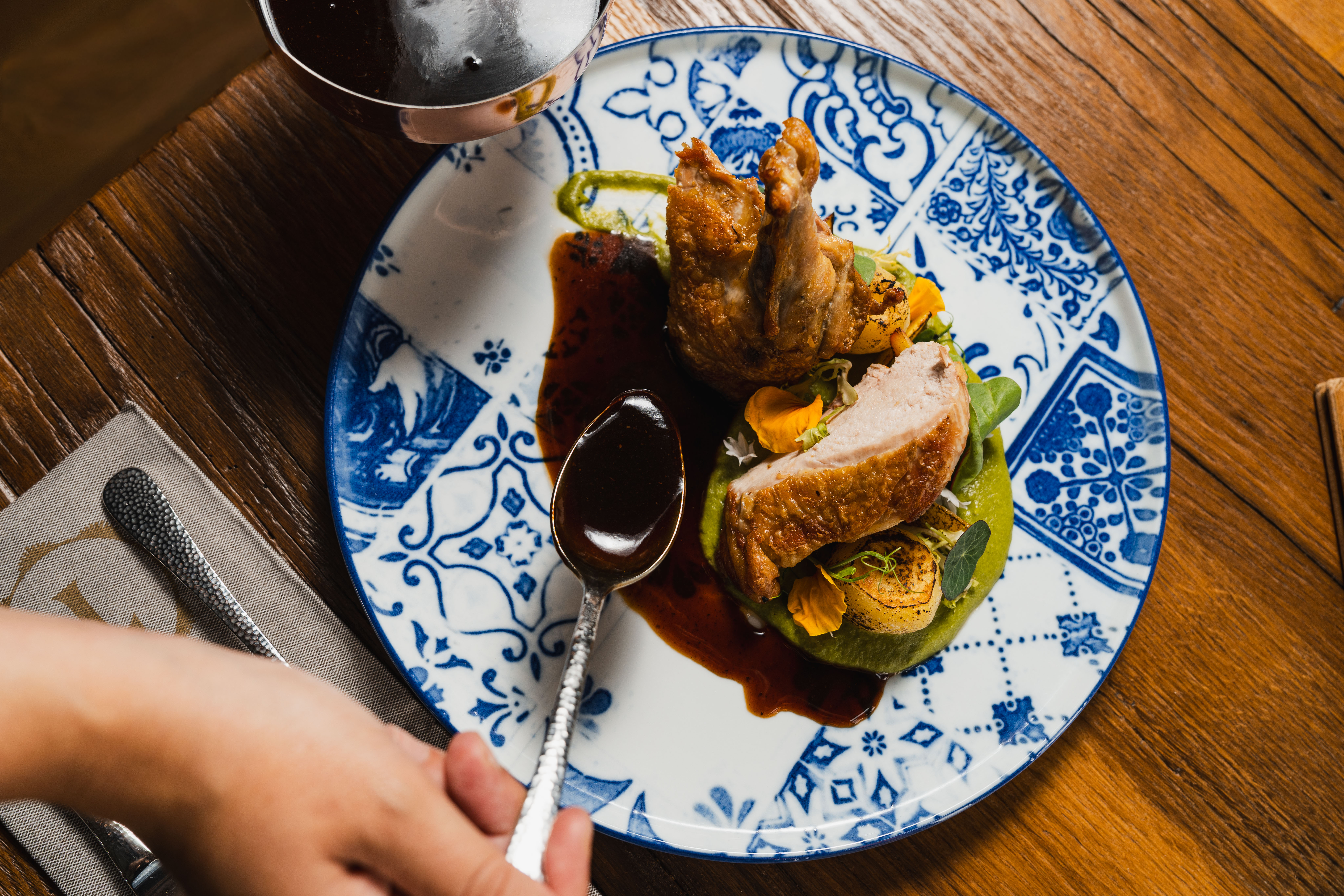 Sauce being poured over a main course on a blue-and-white ceramic plate.